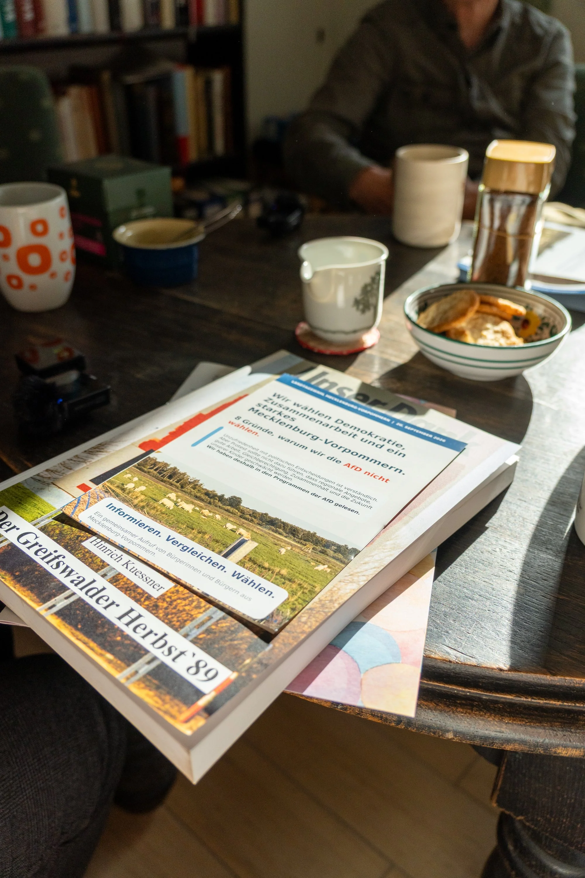 Flyer "8 Gründe warum wir die AfD nicht wählen. - Informieren. Vergleichen. Wählen" auf einem Stapel Bücher auf einem Tisch mit Kaffeetassen.