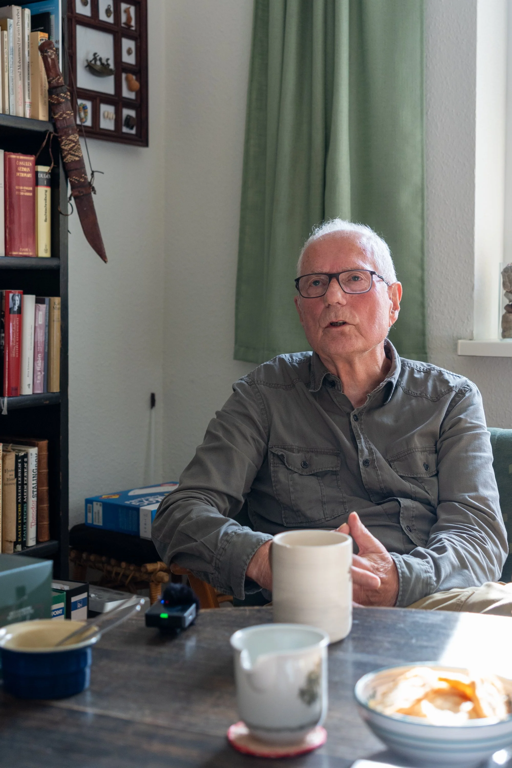 Hinrich Kuessner sitzt an einem Tisch und führt im Raum ein Gespräch, während seine Hände vor einer Tasse gestikulierend zusammengelegt sind.