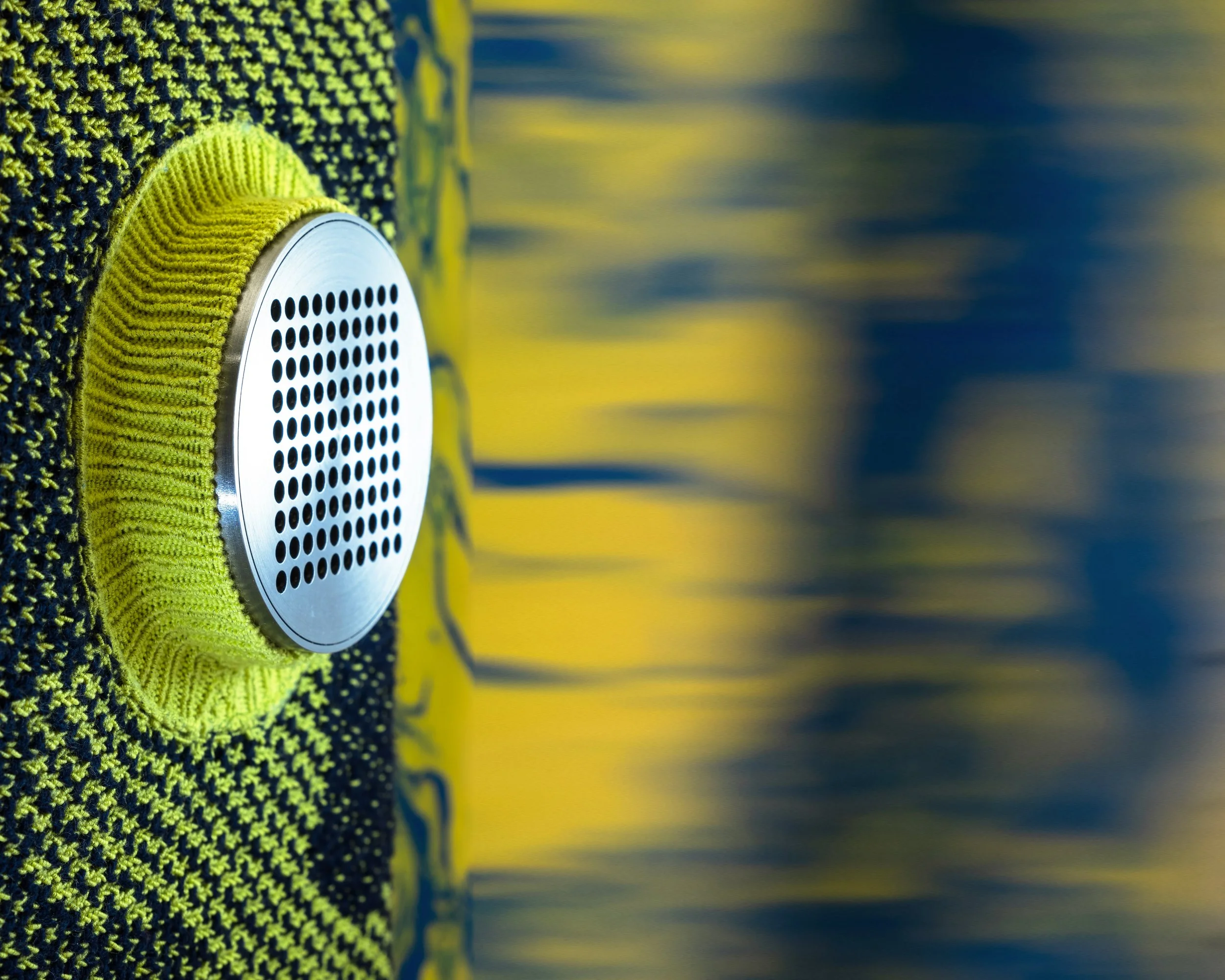 Close-up of a portable speaker with a metallic grille surrounded by yellow fabric, placed on a fabric surface with yellow and black knit pattern, next to a blurred background.