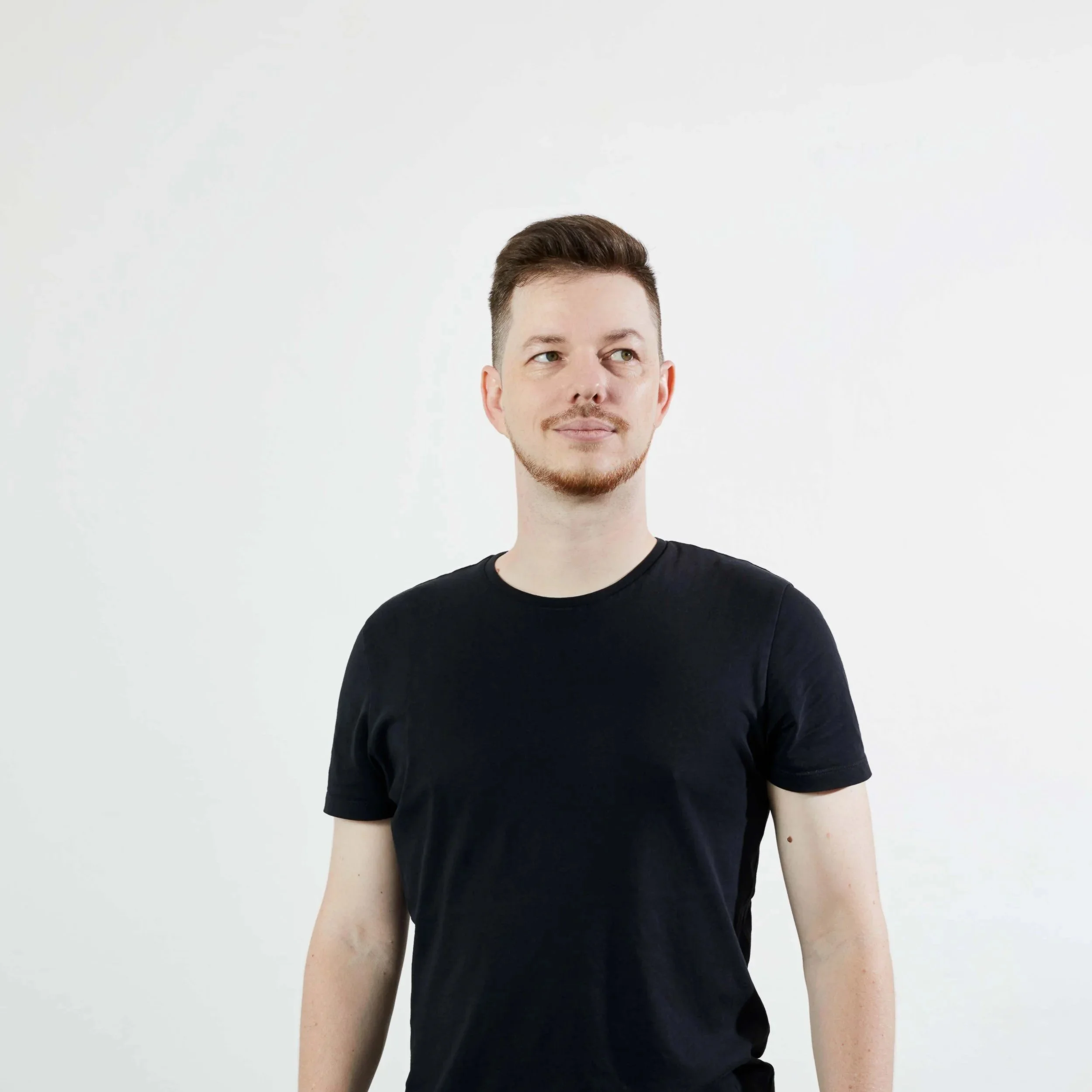 A man with short brown hair and a light beard, wearing a black t-shirt, looks to his left against a plain white background.