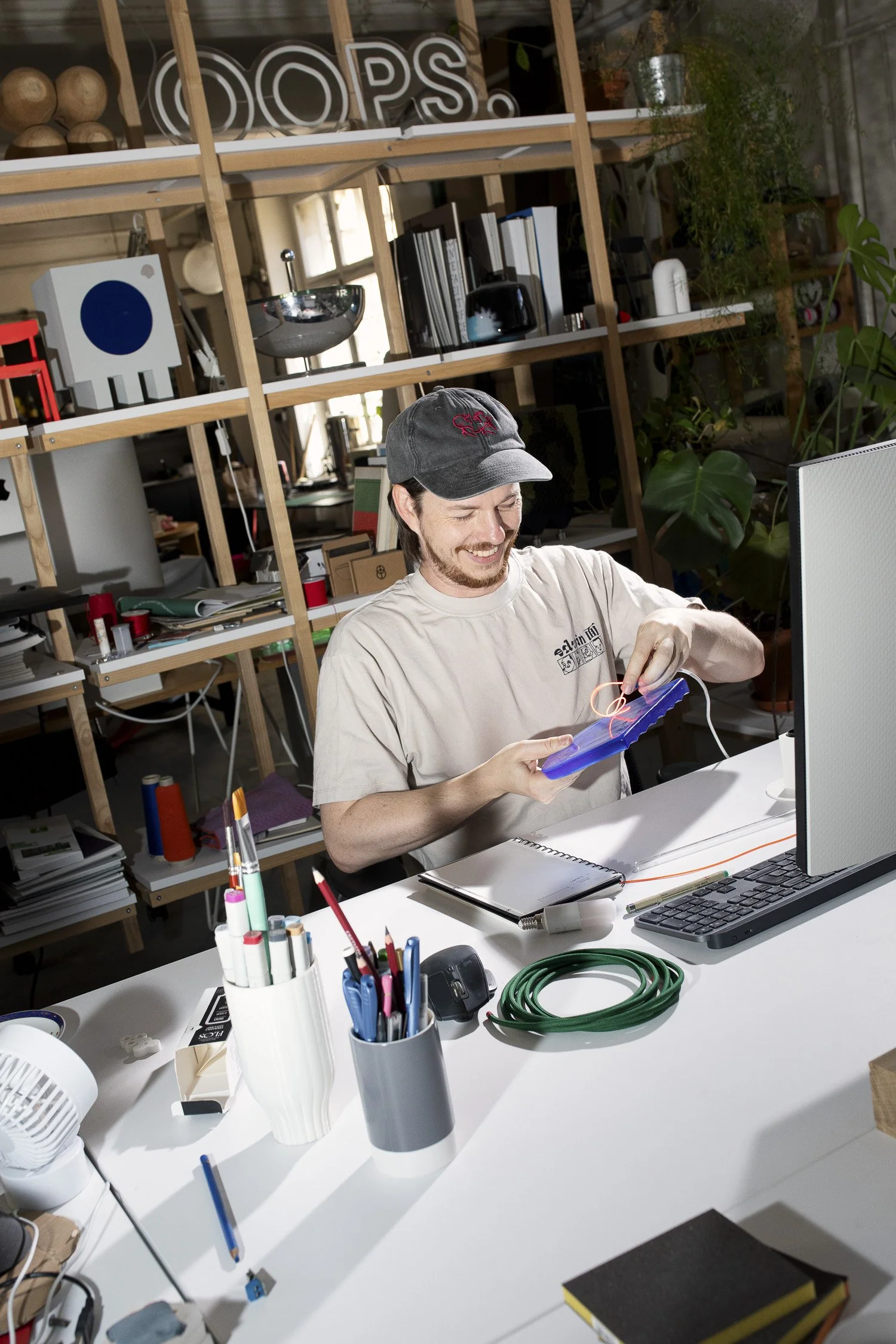 A man in a beige t-shirt and black cap working on an electronics project at a cluttered desk, surrounded by shelves with books, decorative objects, and plants.