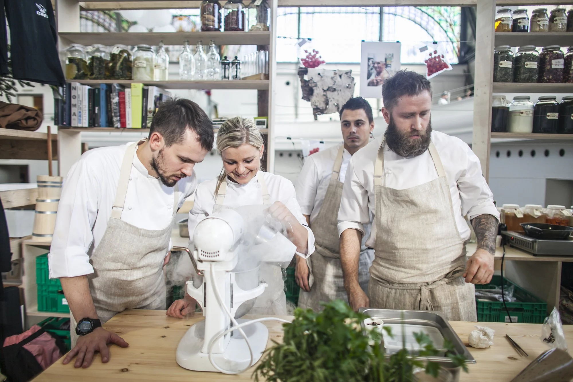 Group of five chefs in white uniforms and aprons working together in a kitchen, with shelves of jars and books behind them, preparing food with a stand mixer and various ingredients on the wooden table.