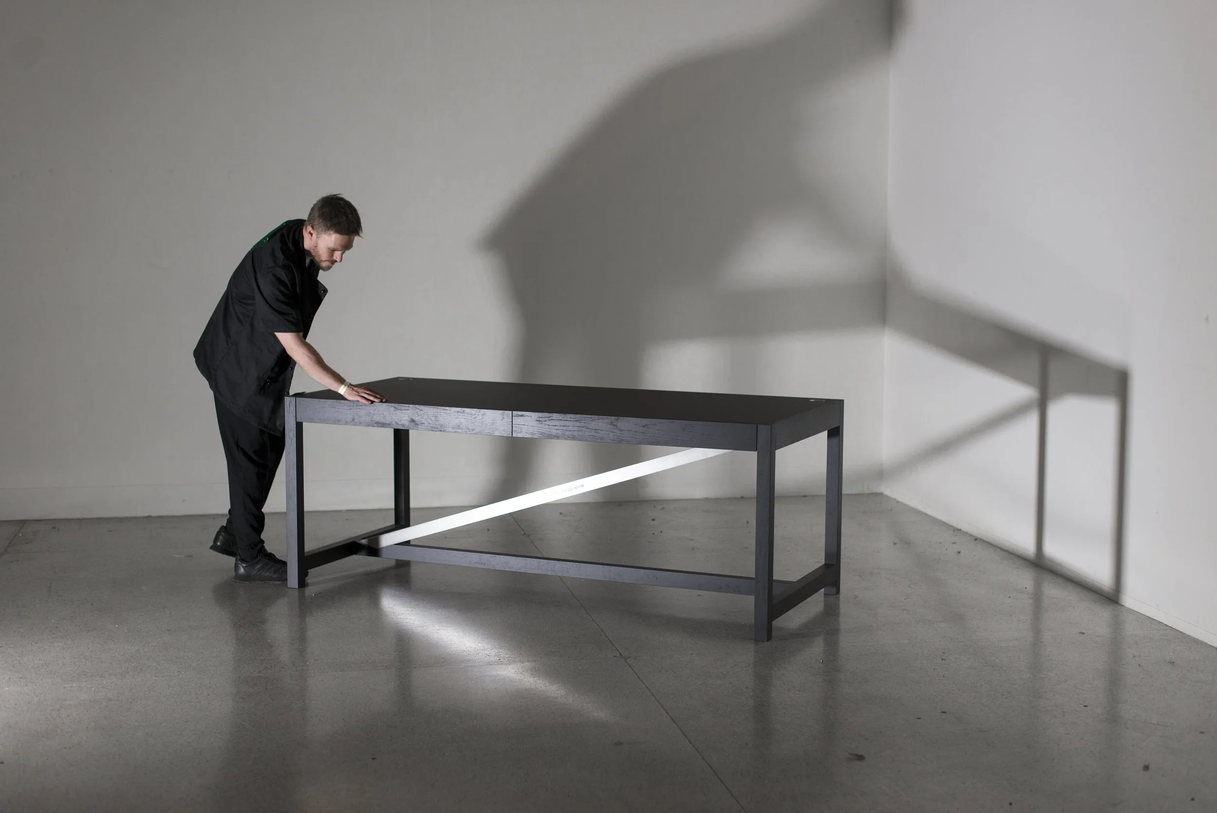 Man in black shirt and pants pushing a black table near a plain white wall in a minimalist setting.