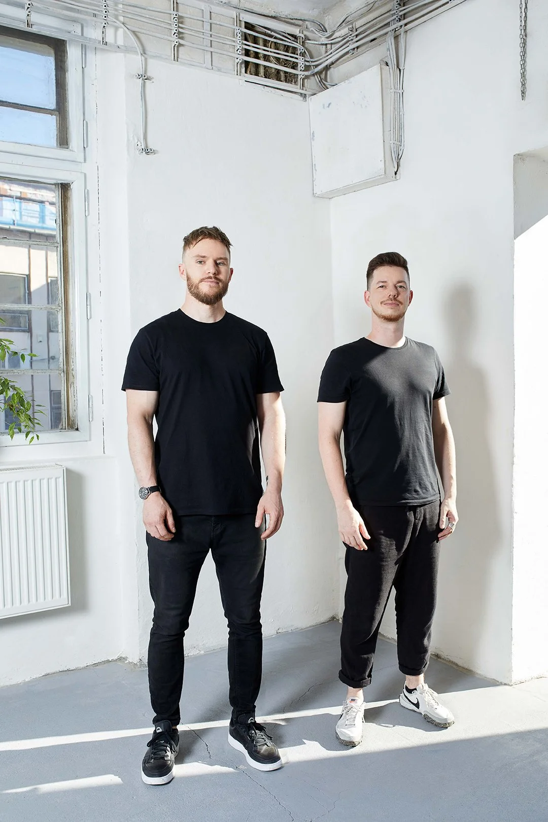 Two young men standing indoors next to each other, both wearing black T-shirts and black pants, with natural light coming from a window on the left.