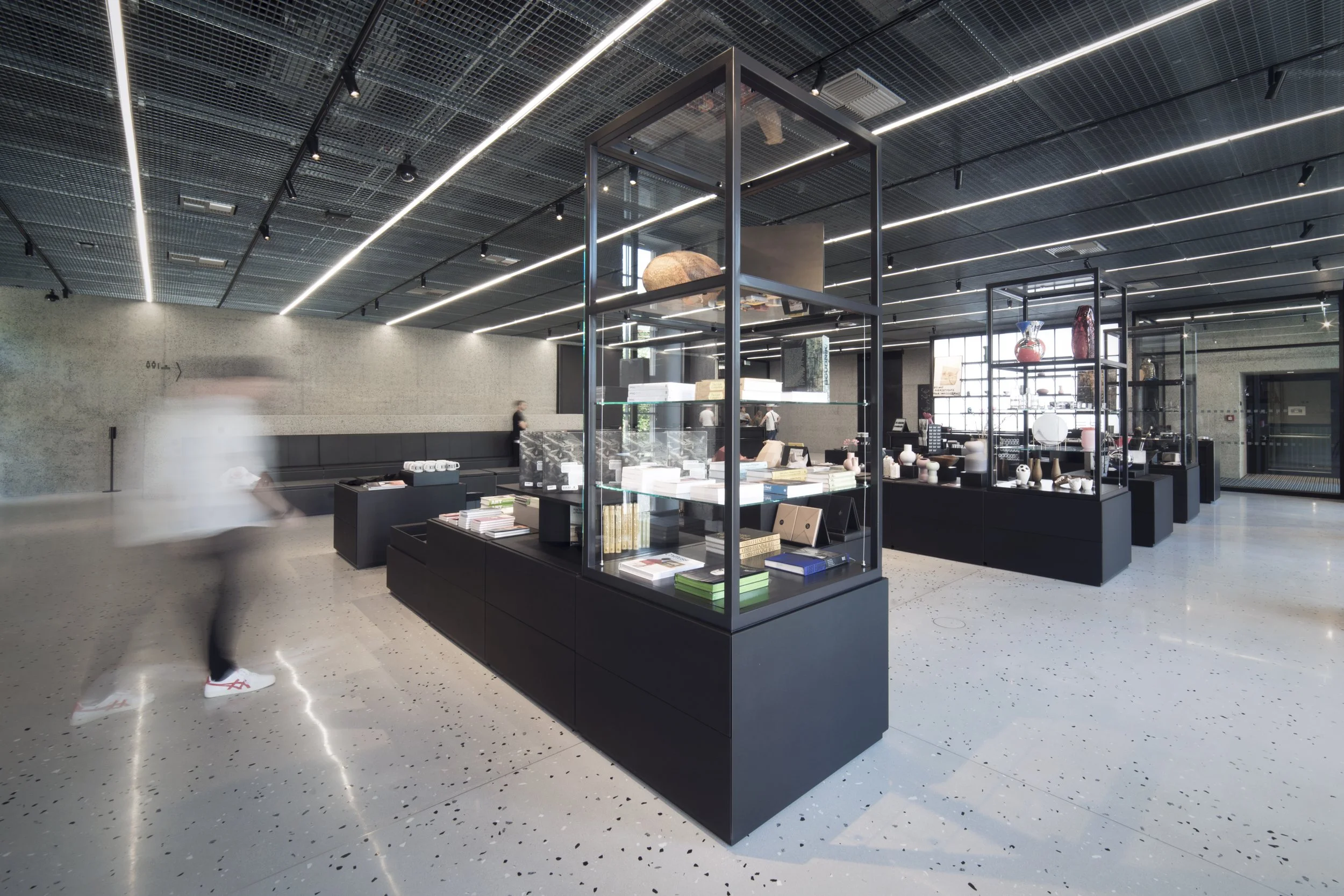 Interior of a modern store or showroom with black display cases filled with books, ceramics, and decorative items, in a spacious area with terrazzo floors, black ceiling with linear lighting, and large windows.