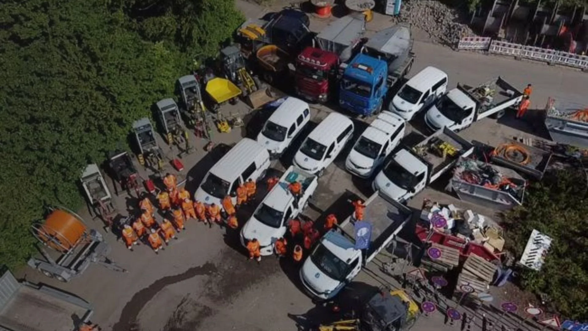 Luftaufnahme eines Baustellenfeldes mit mehreren weißen Fahrzeugen, Bauhelfern in orangefarbener Kleidung, Baumaschinen und Baugeräten.