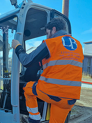 Un trabajador con uniforme naranja y gris sentado en la abertura de una maquinaria, probablemente en una obra en construcción o un sitio industrial, con fondo de edificio y cielo azul.