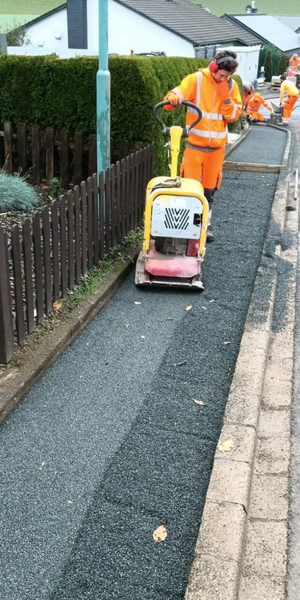 Trabajador de mantenimiento en uniforme naranja colocando asfalto en la acera junto a una cerca de madera y un jardín con setos, en un día soleado.