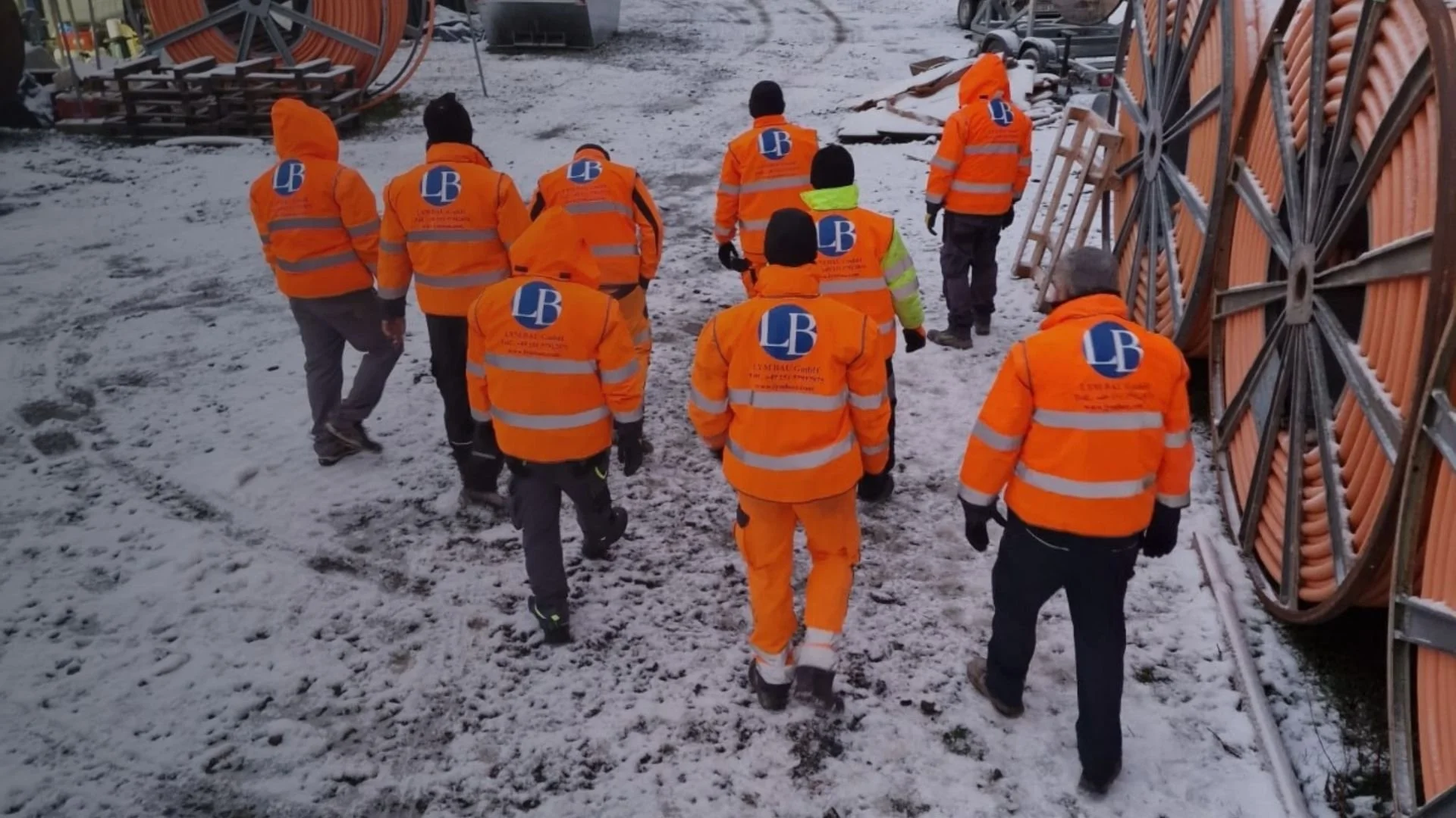 Gruppe von Bauarbeitern in orange Sicherheitskleidung und Mützen, die auf einer Baustelle im Winter gehen, mit Schneebedecktem Boden und Baumaterialien
