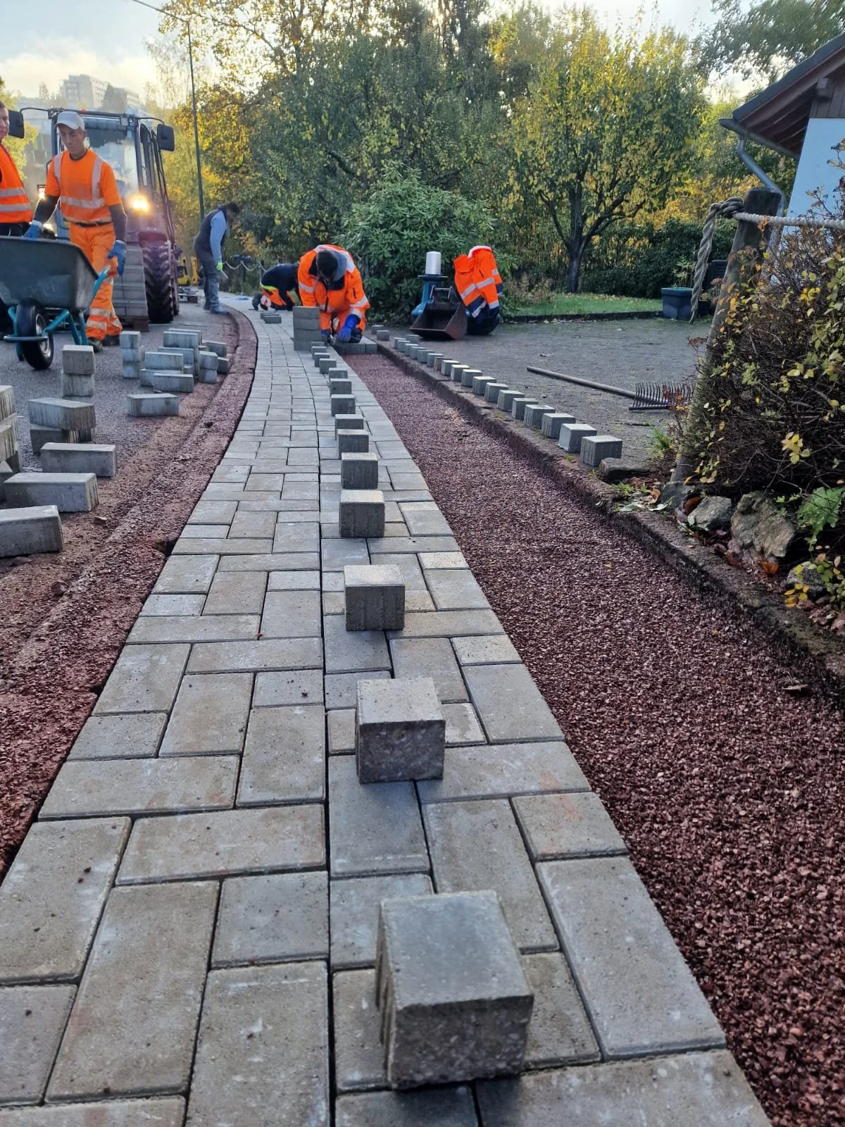 Menschen arbeiten an der Pflasterung eines Gehwegs mit Ziegelsteinen. Sie tragen orange Sicherheitshosen, es ist Tag und es sind Bäume im Hintergrund.