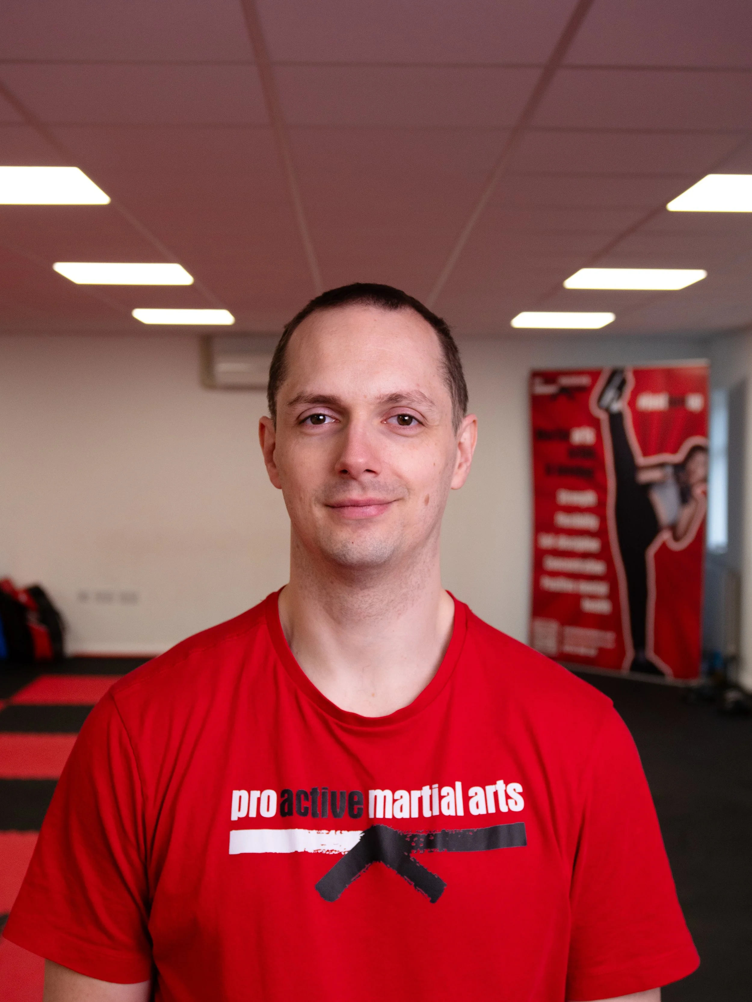 Richard Jago in PAMA red martial arts t-shirt, standing in our martial arts training facility, in Gerrards Cross with mats and a poster of a person performing a high kick in the background.