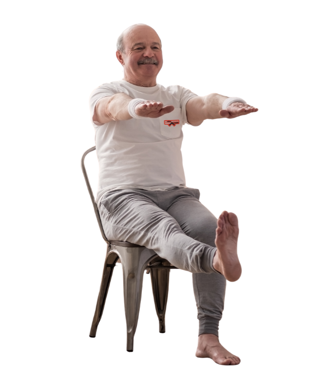 An elderly Healthspan Heros man with a mustache, wearing a white t-shirt and gray sweatpants, sitting on a metal chair with one arm extended forward and one leg lifted, smiling.