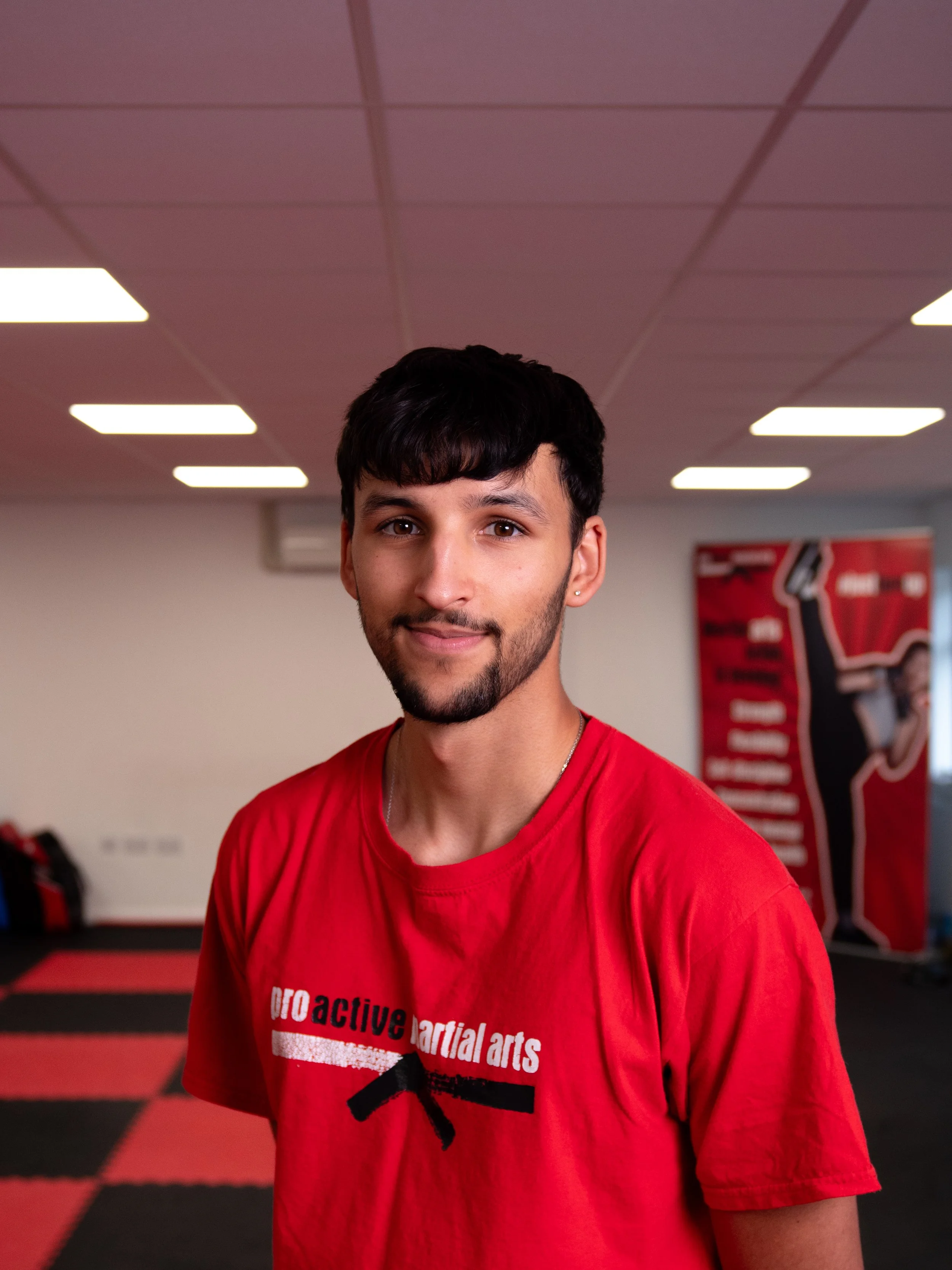 Young man with dark hair and a beard wearing a red T-shirt with martial arts logo inside a martial arts studio.
