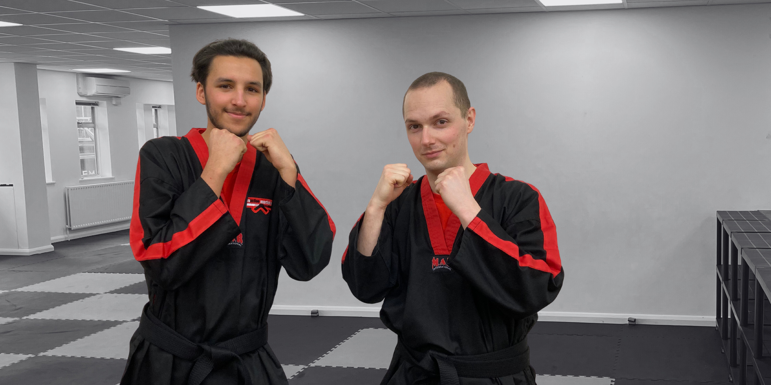 Two men dressed in black and red martial arts uniforms in a training dojo, standing in fighting stances with fists raised.