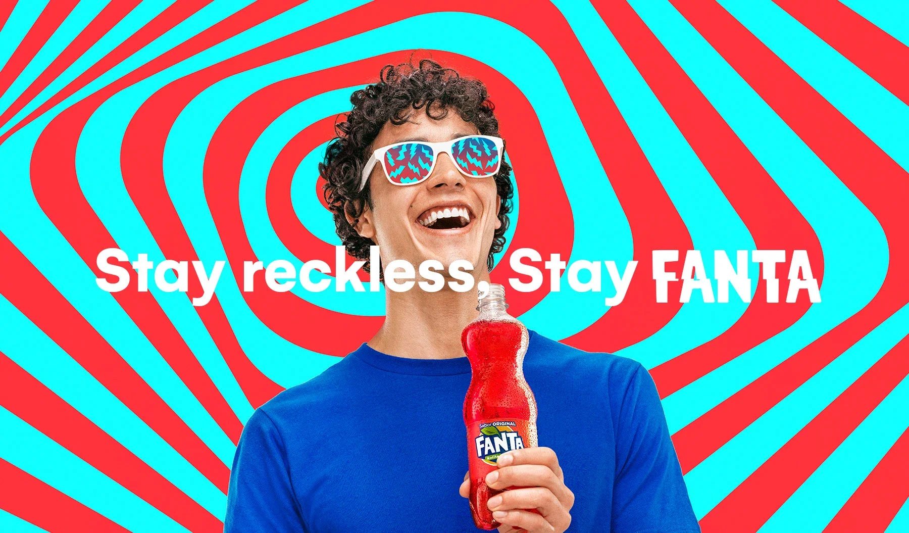 A smiling person wearing colorful sunglasses and a blue shirt, holding a Fanta soda bottle, with a red and blue swirling background and the slogan 'Stay reckless, Stay FANTA' in white and red text.