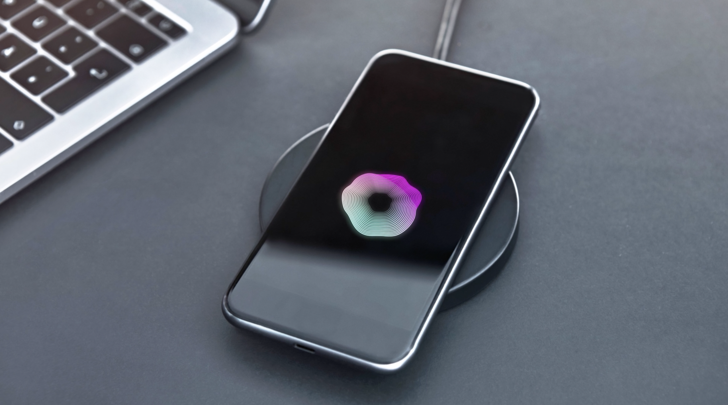 Smartphone on wireless charger with colorful holographic donut-shaped graphic on screen, next to a laptop on a gray surface.