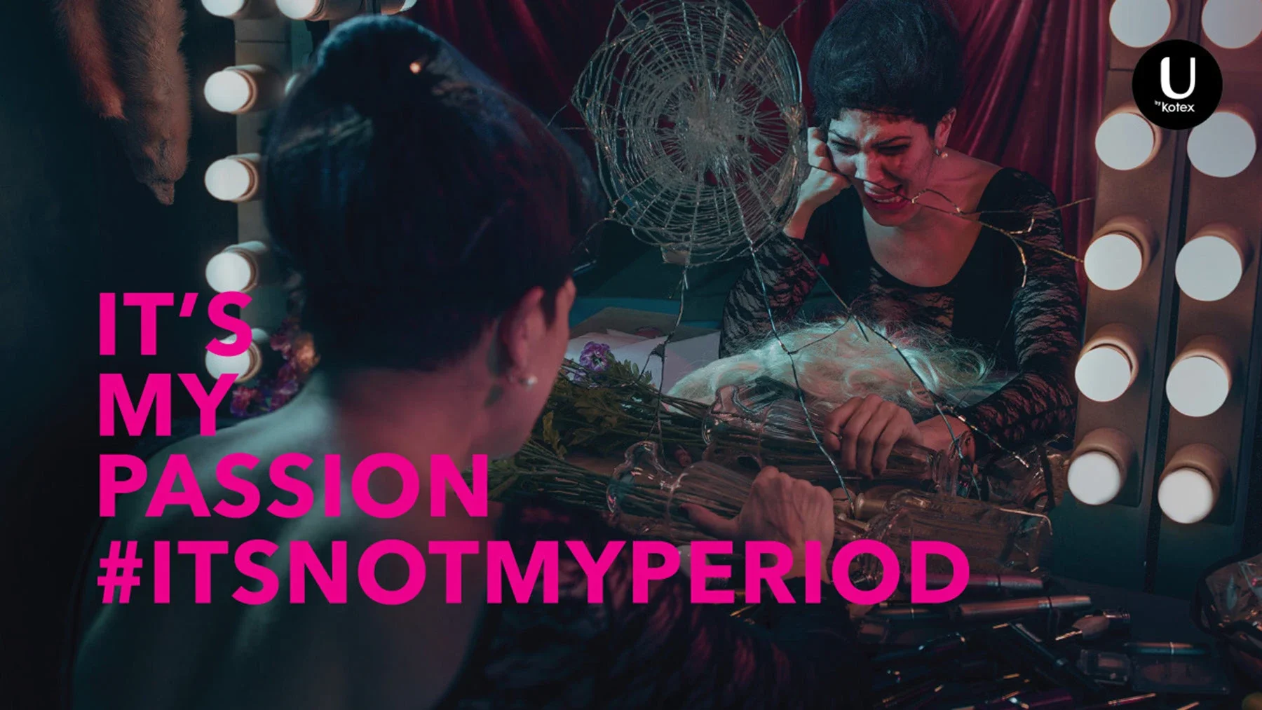A woman smiling at a mirror with lights, holding a bouquet of flowers, with another person facing her. The scene suggests a makeup or dressing room. Text overlay: 'IT'S MY PASSION #ITSNOTMYPERIOD' in pink.