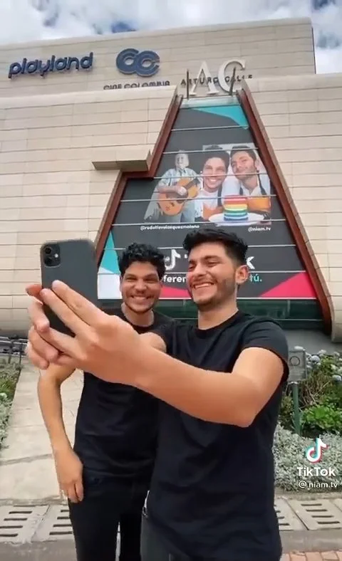 Two young men in black shirts taking a selfie in front of a building with a large sign showing two people with guitars and colorful objects, with a building labeled 'Playland' and 'Oceana' in the background.