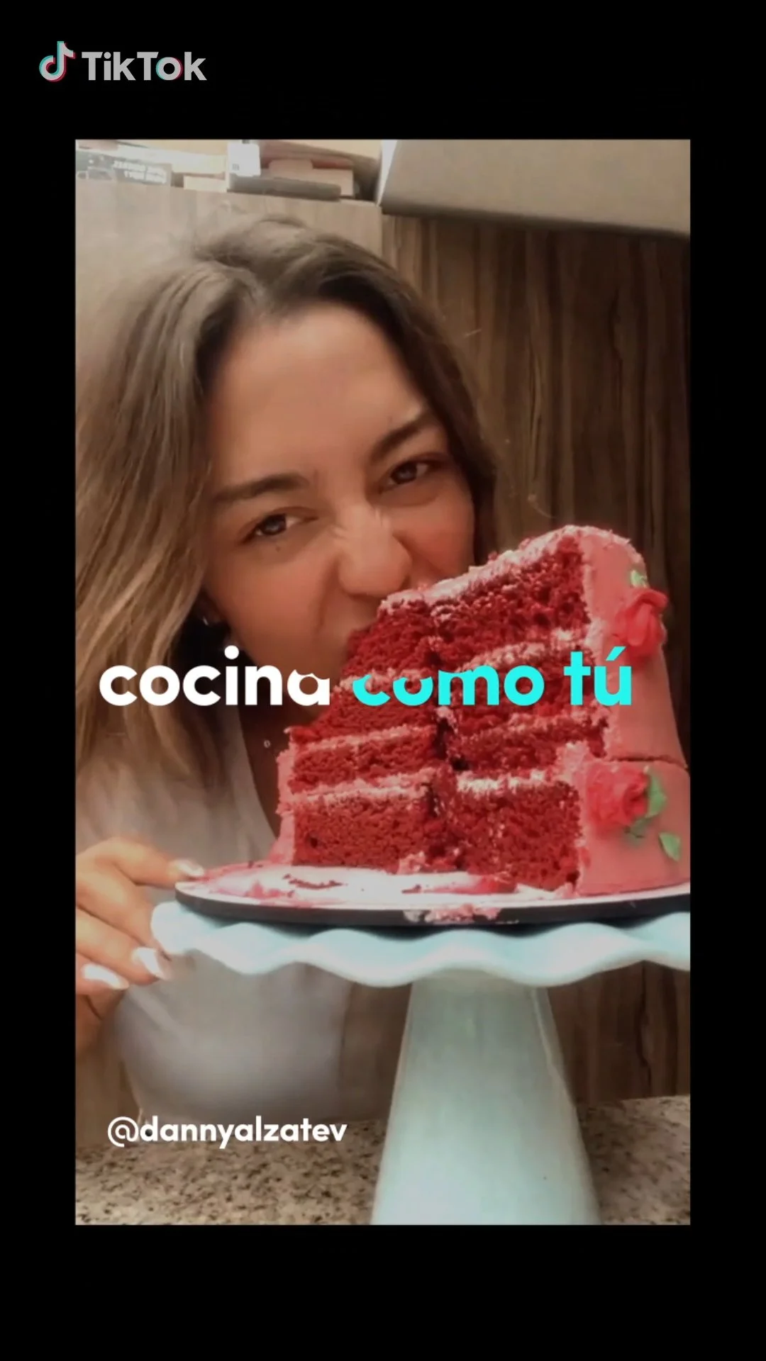 A young woman with light brown hair taking a bite of a large red velvet cake with pink icing and floral decorations on a white cake stand.