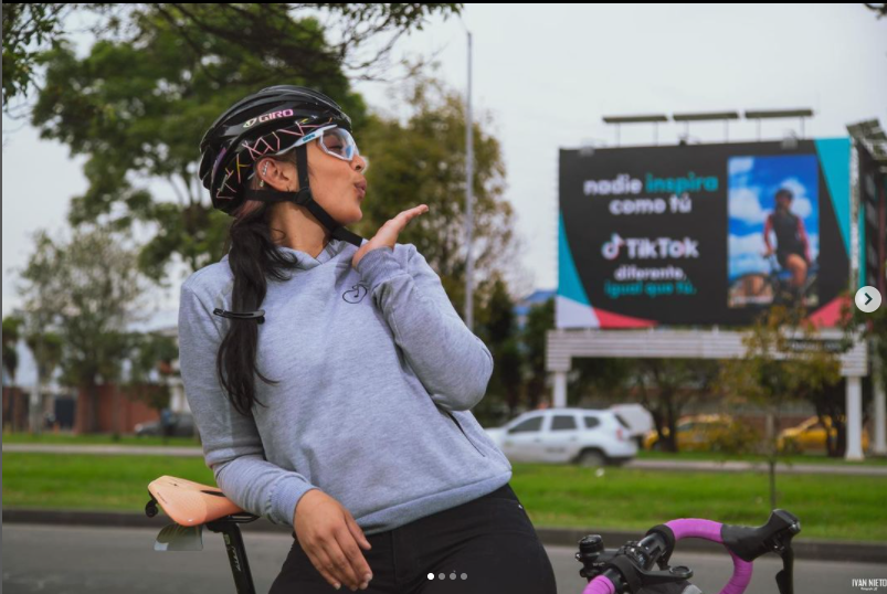 A woman wearing a helmet and sunglasses making a kissy face gesture outdoors, standing next to a bicycle with a billboard in the background.
