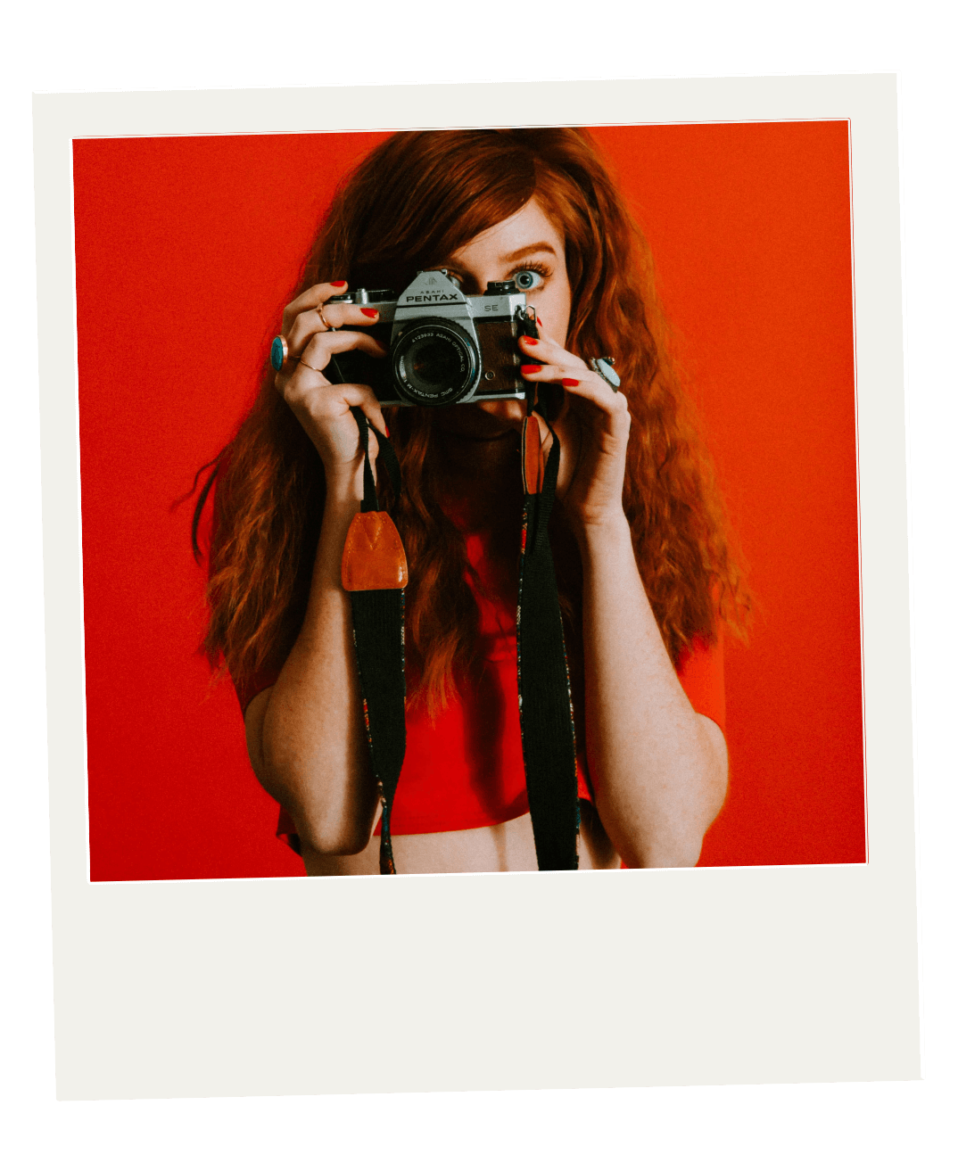 A woman with long red hair, wearing a red top, takes a photo with a vintage camera against a red background. She is holding the camera to her face with both hands, and the camera strap is hanging down.