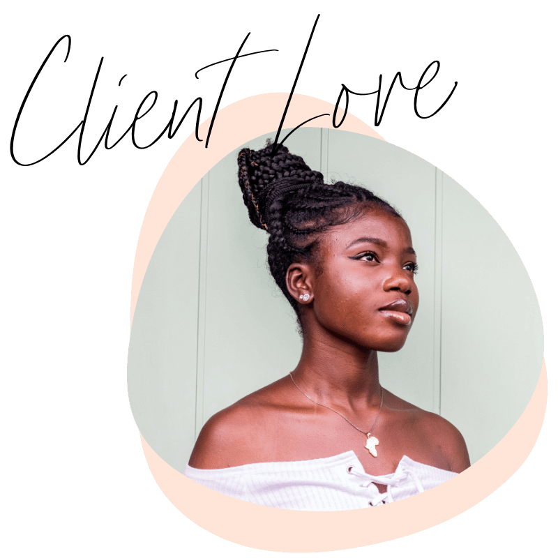 Portrait of a young African American woman with braided hair styled in an updo, wearing an off-shoulder white top, a necklace with the shape of Africa, stud earrings, standing against a light-colored background.