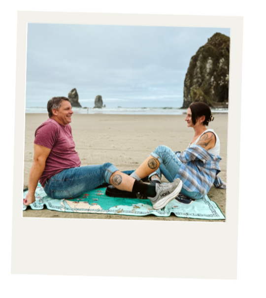 Two people sitting on a beach blanket facing each other, smiling, with large rock formations in the background during daytime.