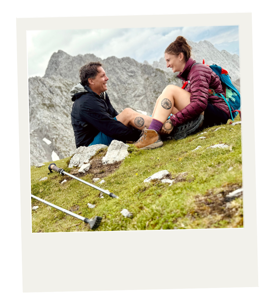 A man and woman sitting on a grassy hillside in mountain terrain, facing each other and smiling, with hiking poles on the ground nearby, mountain peaks in the background.