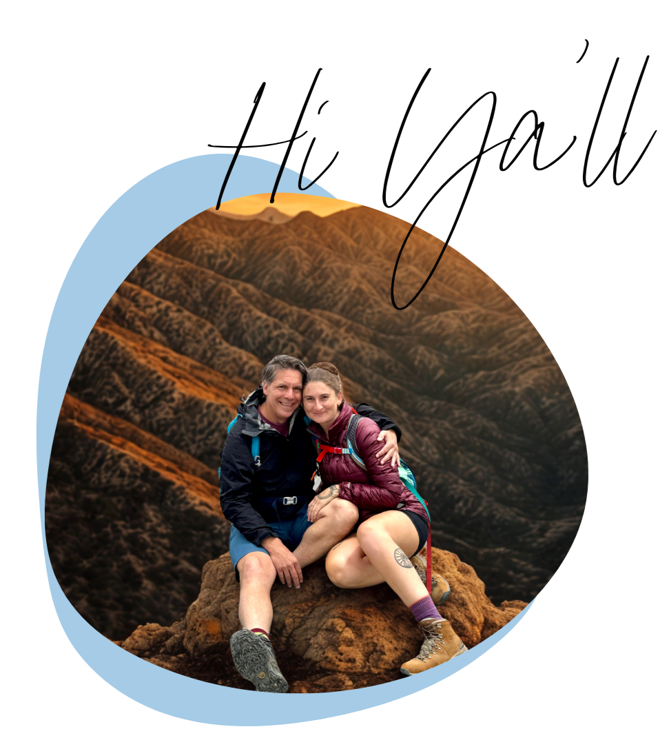 Two hikers sitting on a rock, hugging, with a mountain range in the background and a clear sky.
