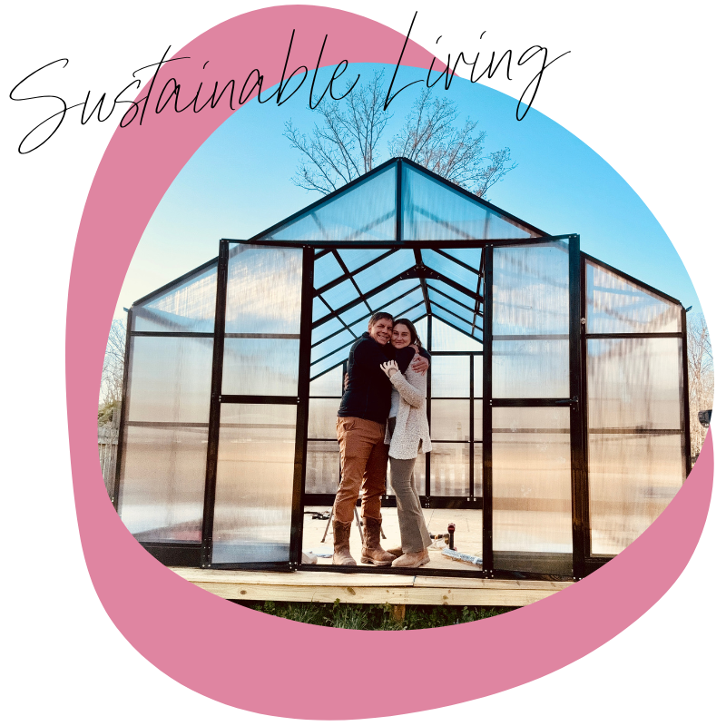 Two women standing inside a small, glass greenhouse, hugging each other, with a tree and blue sky in the background.