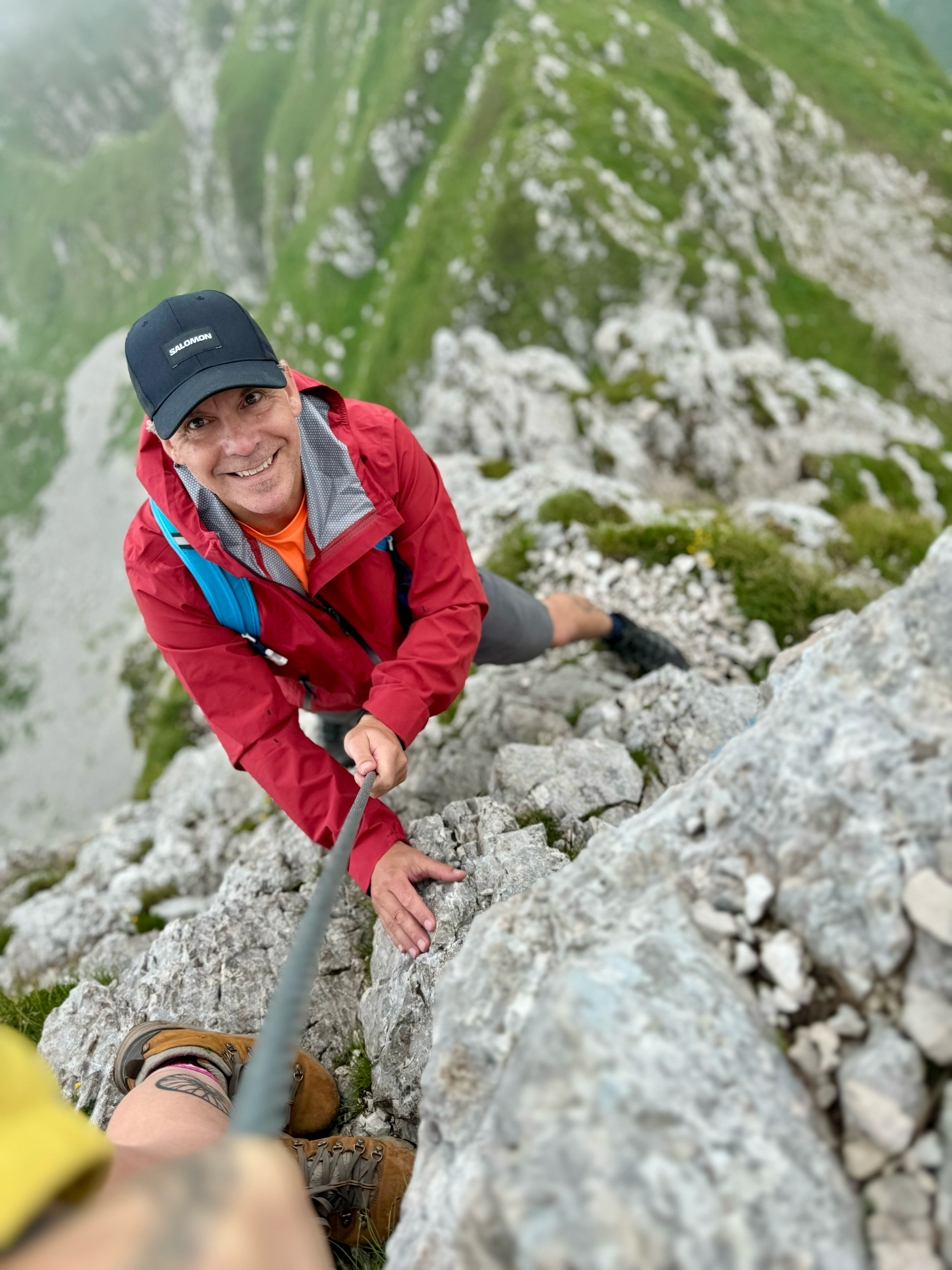 Climber ascending a rocky mountain face, smiling at the camera, wearing a black cap, red jacket, and brown hiking boots.