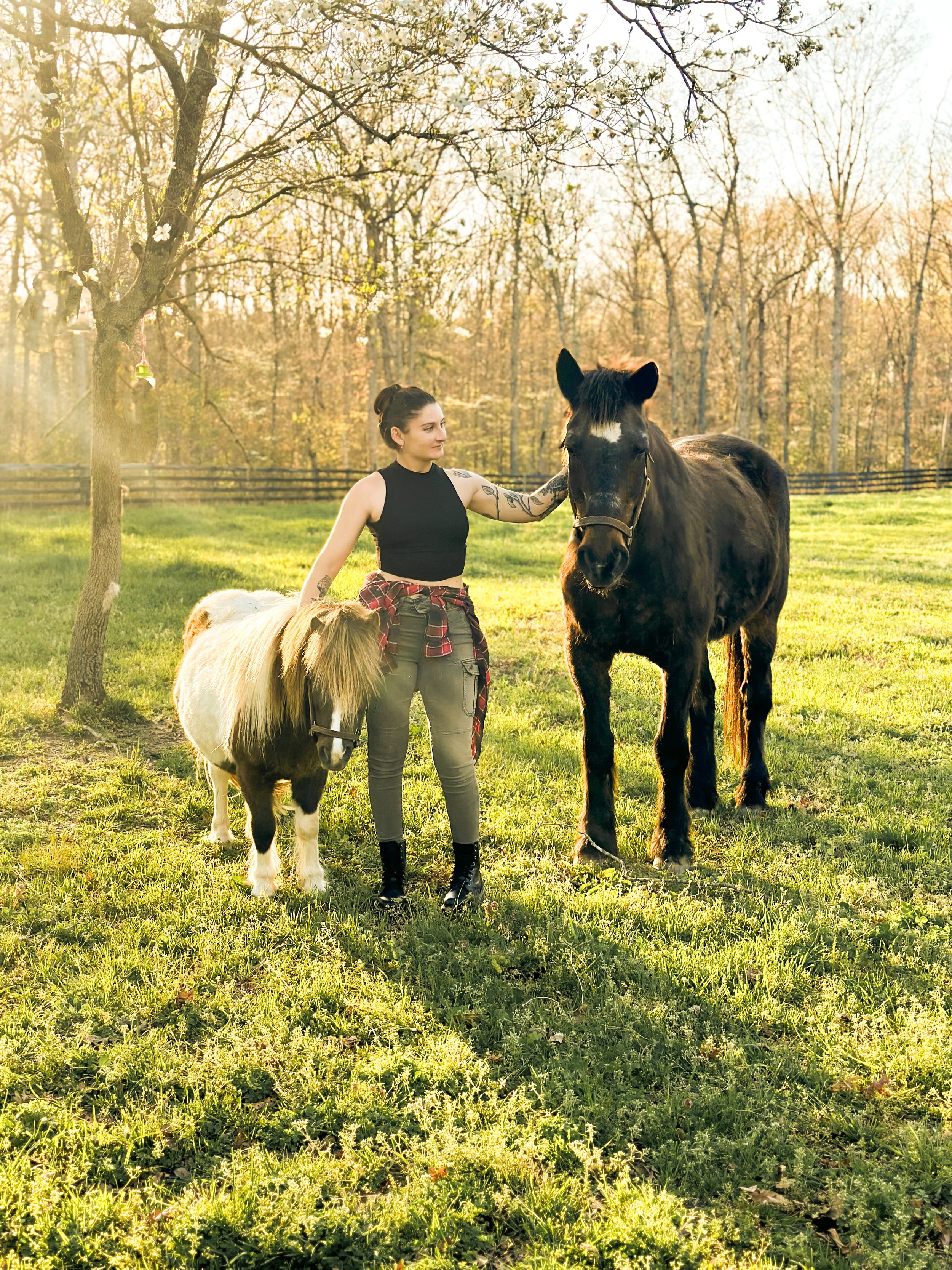 A woman with tattoos, dressed in a black sleeveless top and gray pants, standing in a grassy field with two horses, one smaller with a light-colored mane and the other larger with a dark mane, during sunset in autumn with bare trees in the background.