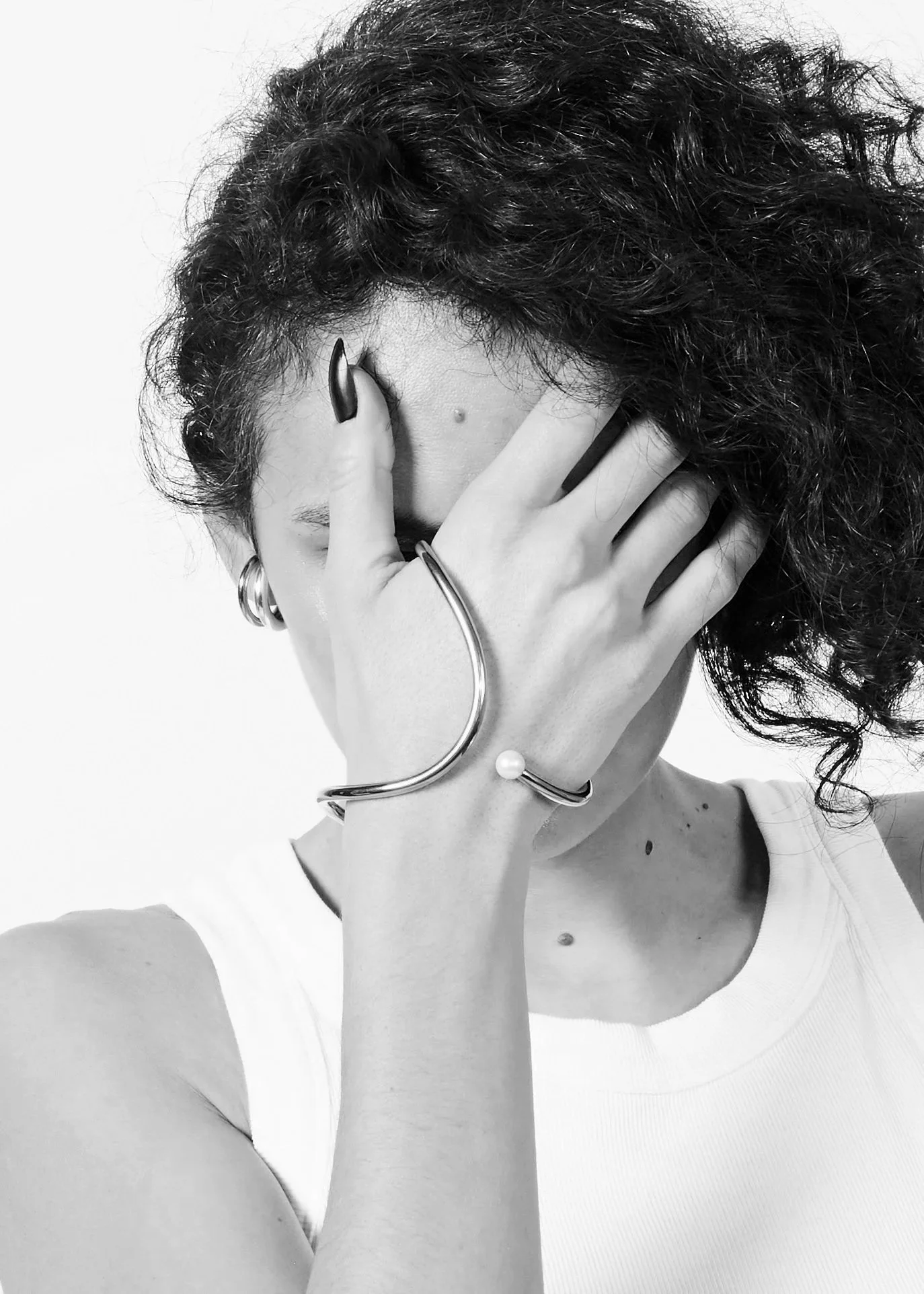 Black and white photo of a woman with curly hair covering part of her face with her hand, wearing hoop earrings and a bracelet with a pearl, dressed in a sleeveless top.