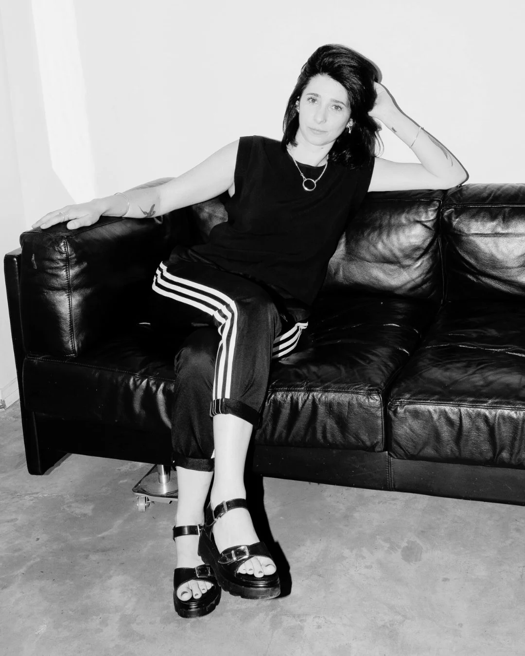 A woman with black hair and tattoos on her arms sitting on a black leather sofa, wearing black sandals, a black sleeveless top, and black athletic pants with white stripes, posing with one arm resting on the sofa and the other hand in her hair.