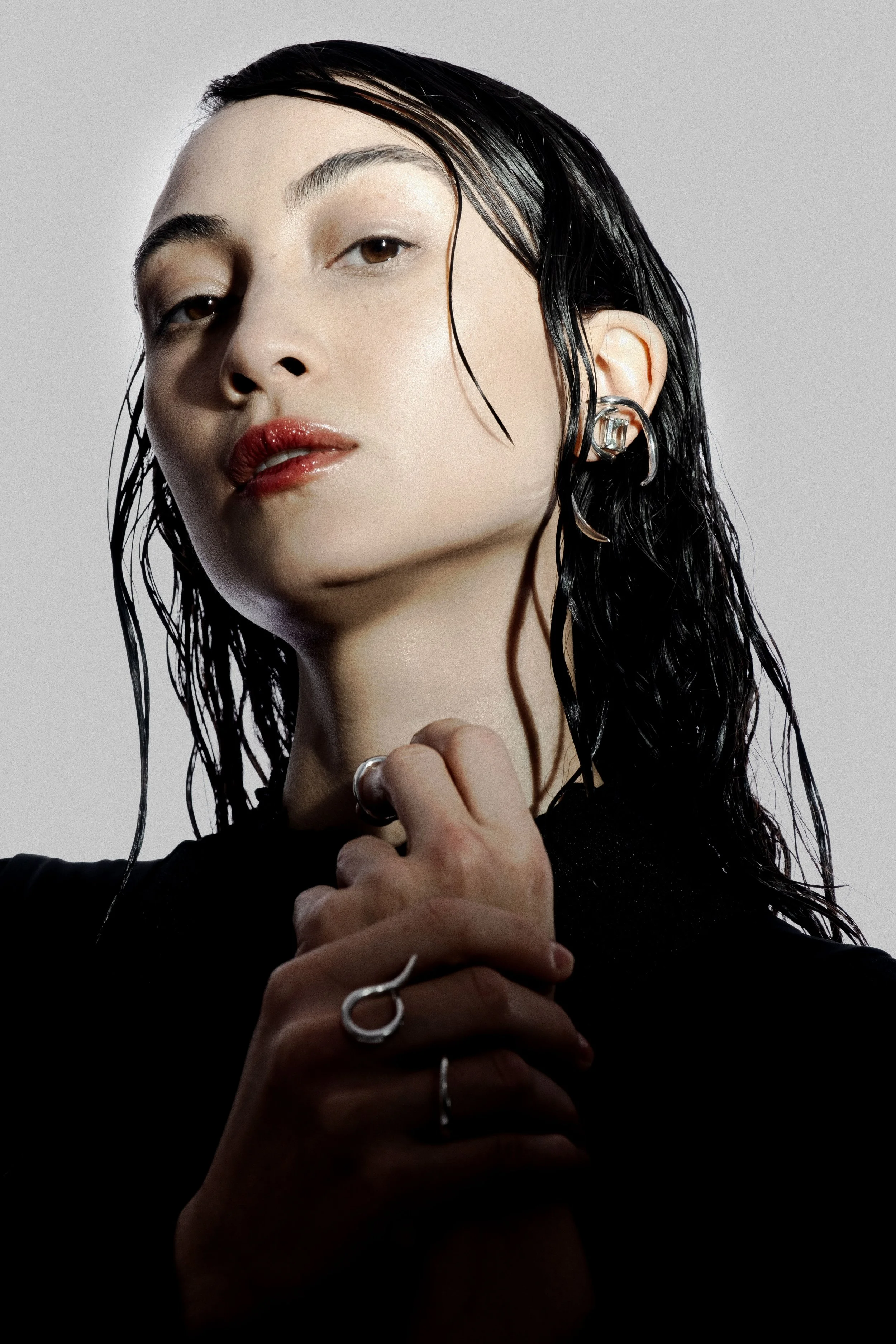 Close-up of a woman with wet black hair wearing earrings and rings, against a plain light background.