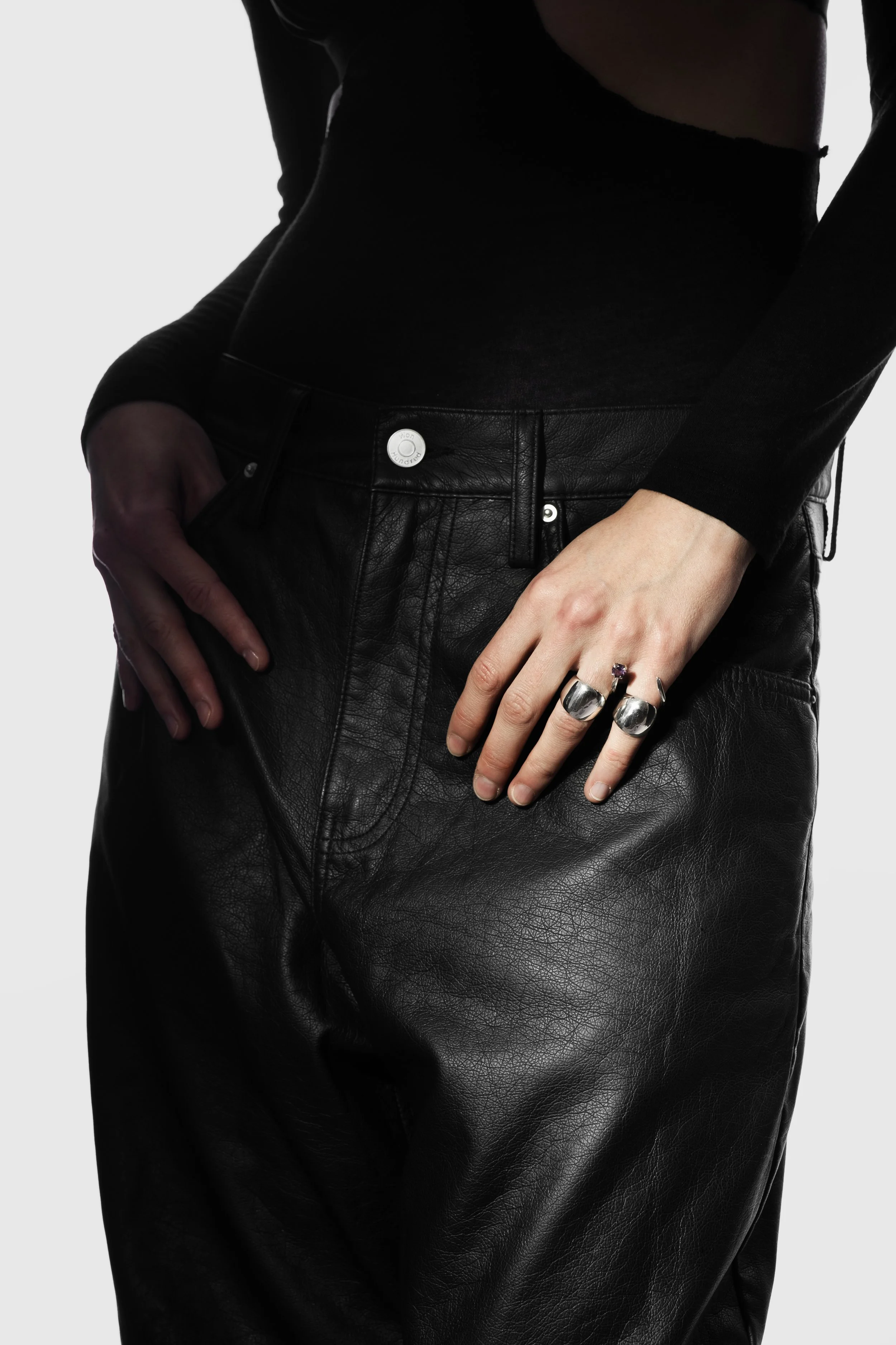 Close-up of a person wearing black leather pants and a black long-sleeve top, with hands resting on hips and wearing rings.