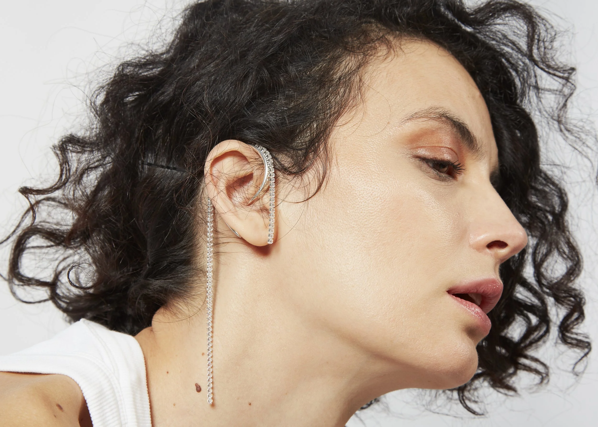 Close-up of a woman's face with curly hair and a long, dangling diamond earring, wearing a white top.