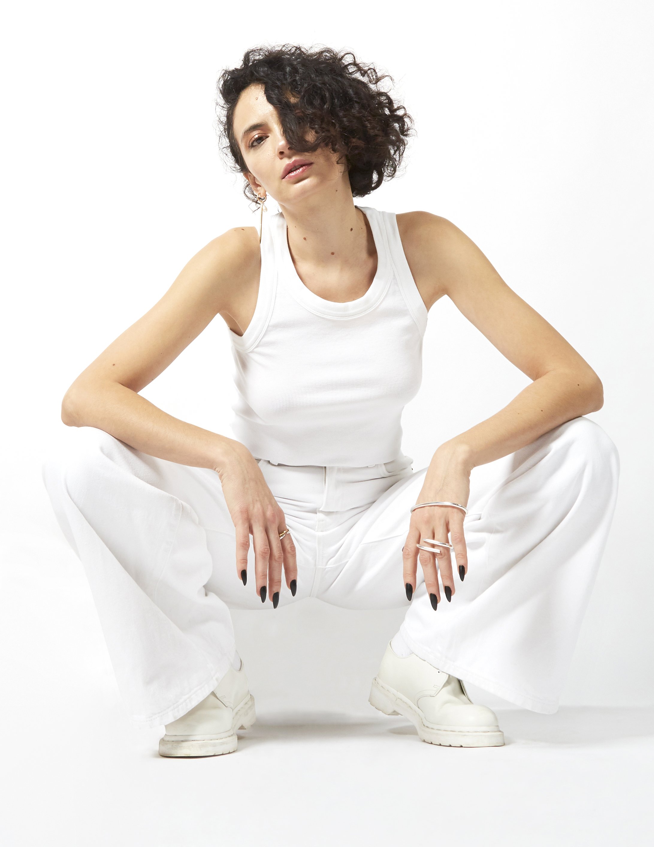 A woman with curly hair wearing white tank top, white wide-leg pants, and white boots, sitting in a squatting pose against a plain white background.
