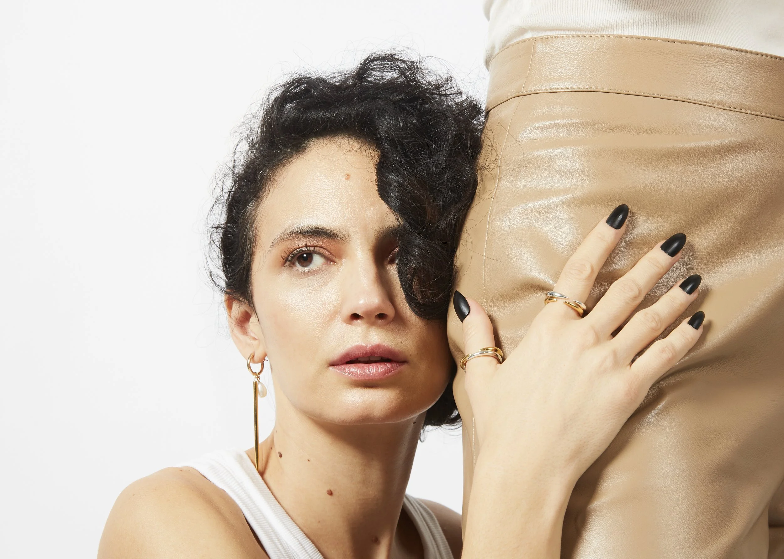 A woman with dark, curly hair and light skin, wearing gold jewelry, leans her head against a person's waist wearing beige leather pants, and has her hand resting on the person's hip.