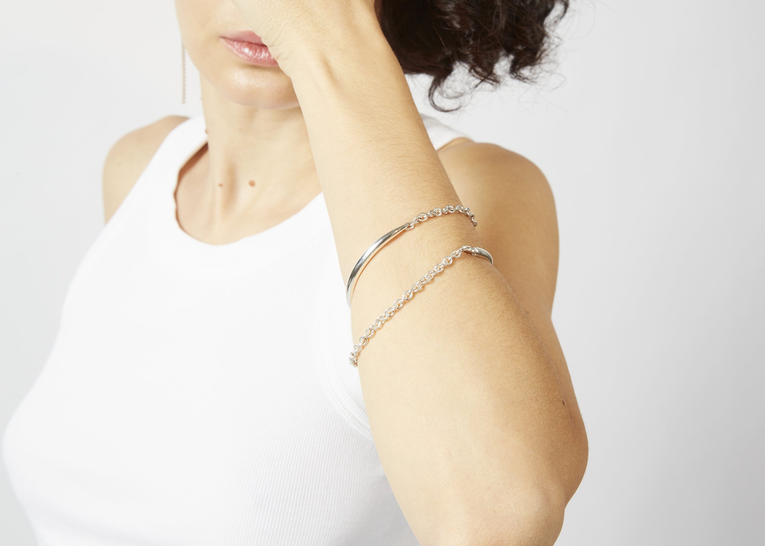 A woman wearing a white sleeveless top and rings on her wrist, with her face partially obscured by her hand, against a plain light background.