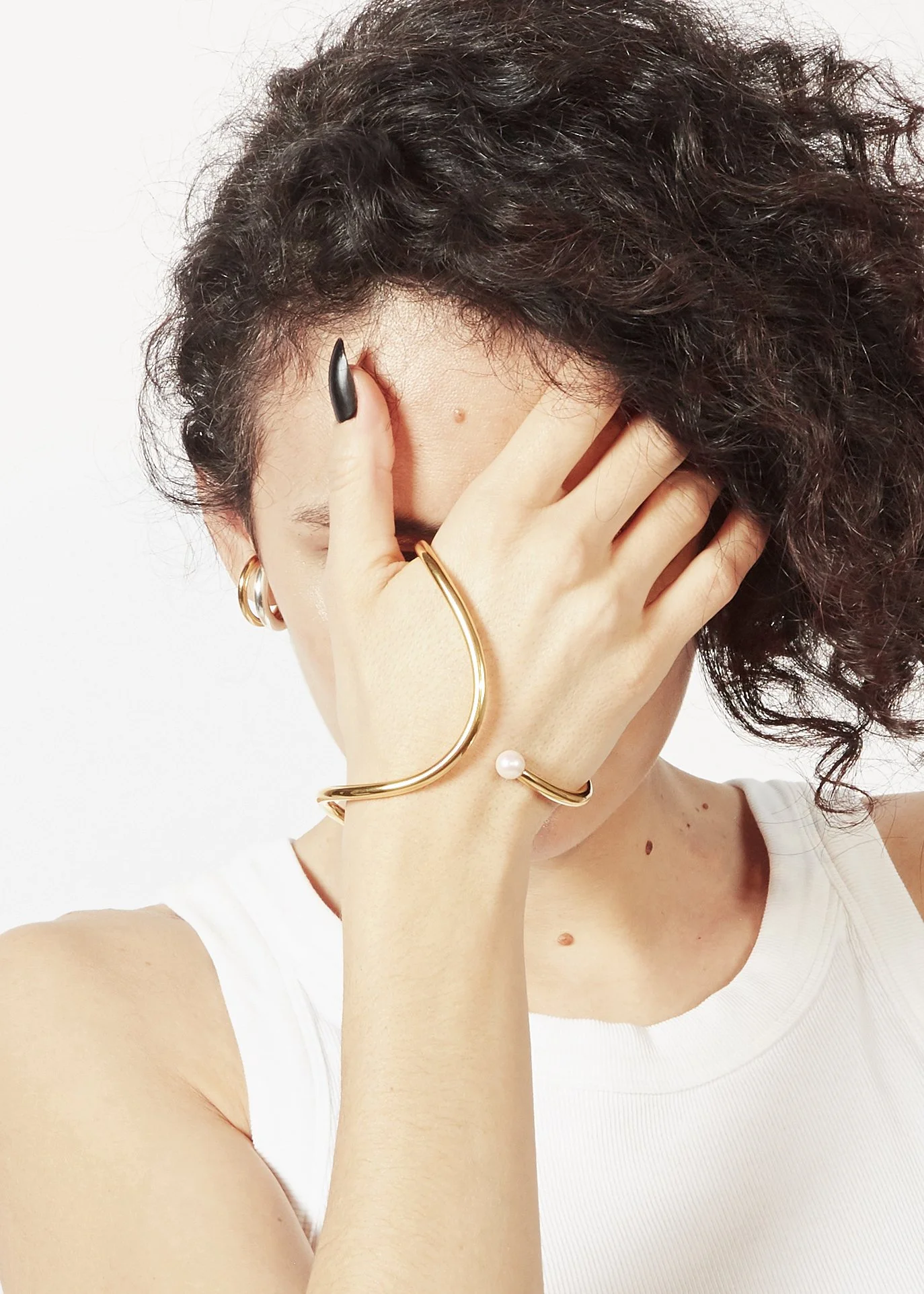 A woman with curly hair covering her face with her hand, showing gold jewelry including a bracelet with a pearl, rings, and earrings, and wearing a white sleeveless top.