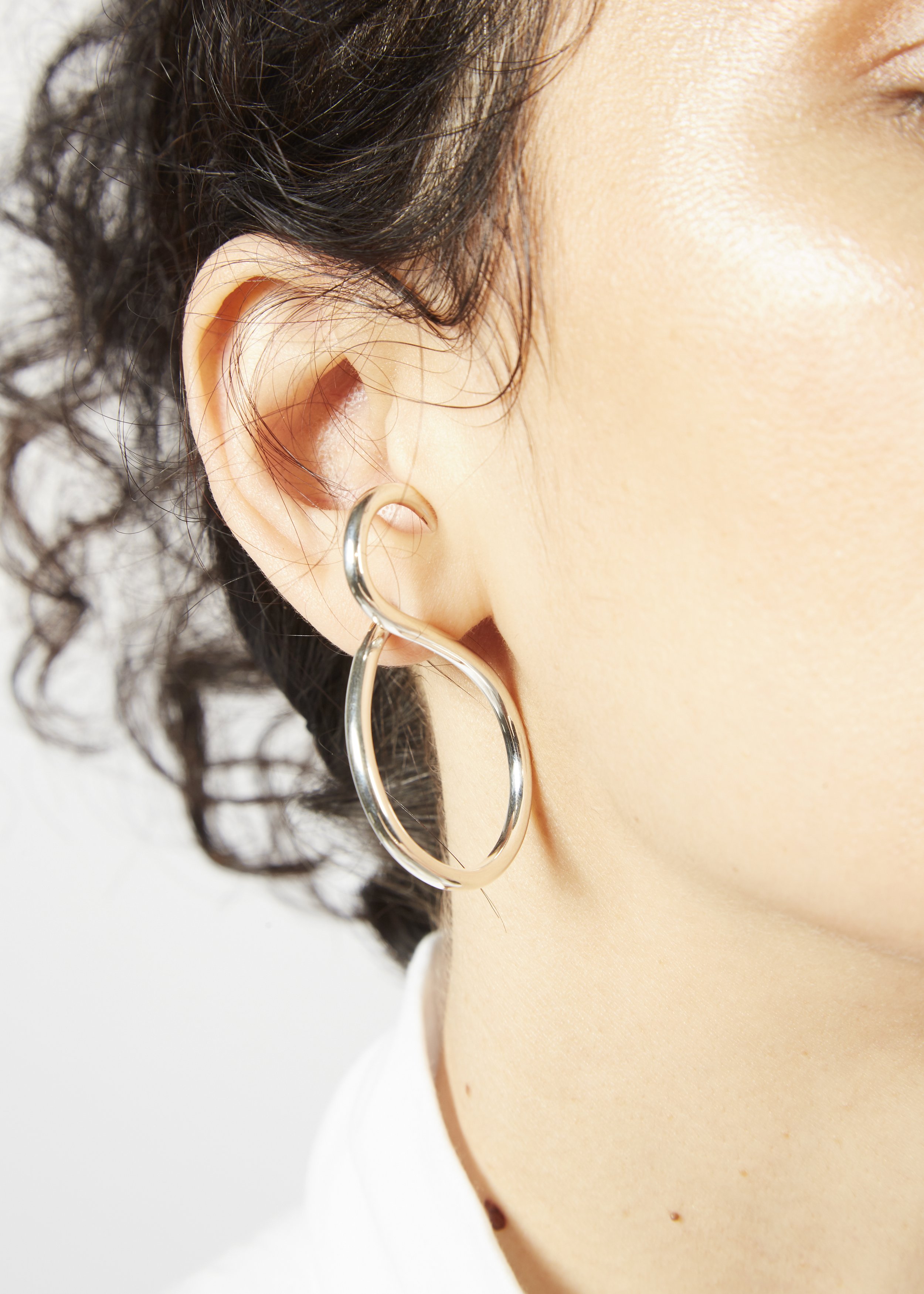 Close-up of a woman's ear with a silver hoop earring, curly dark hair, and a portion of her face visible.