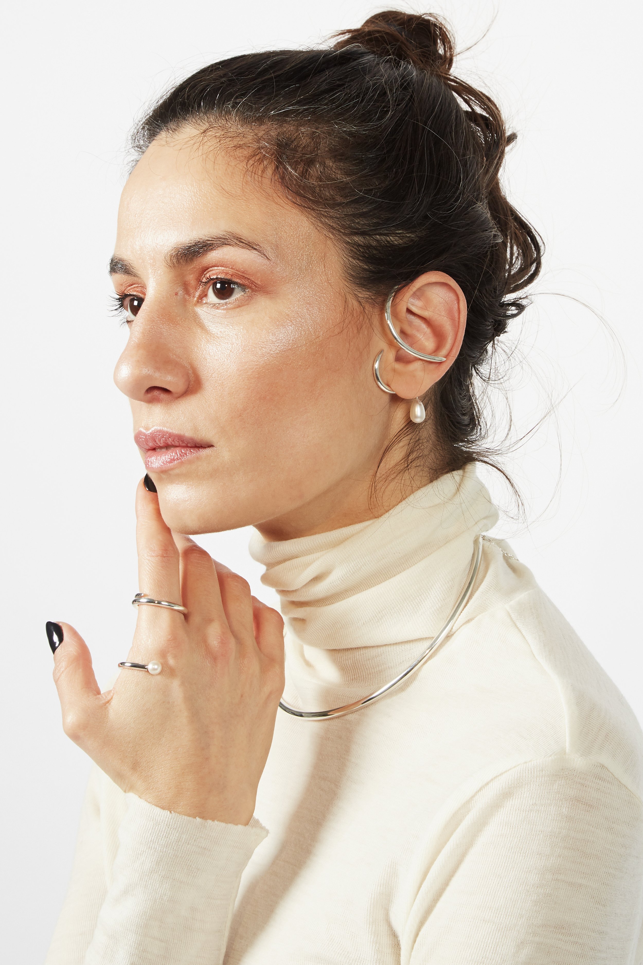 A woman with dark hair styled in an updo, wearing pearl earrings, a silver necklace, and multiple rings, is touching her chin with her left hand against a plain white background.