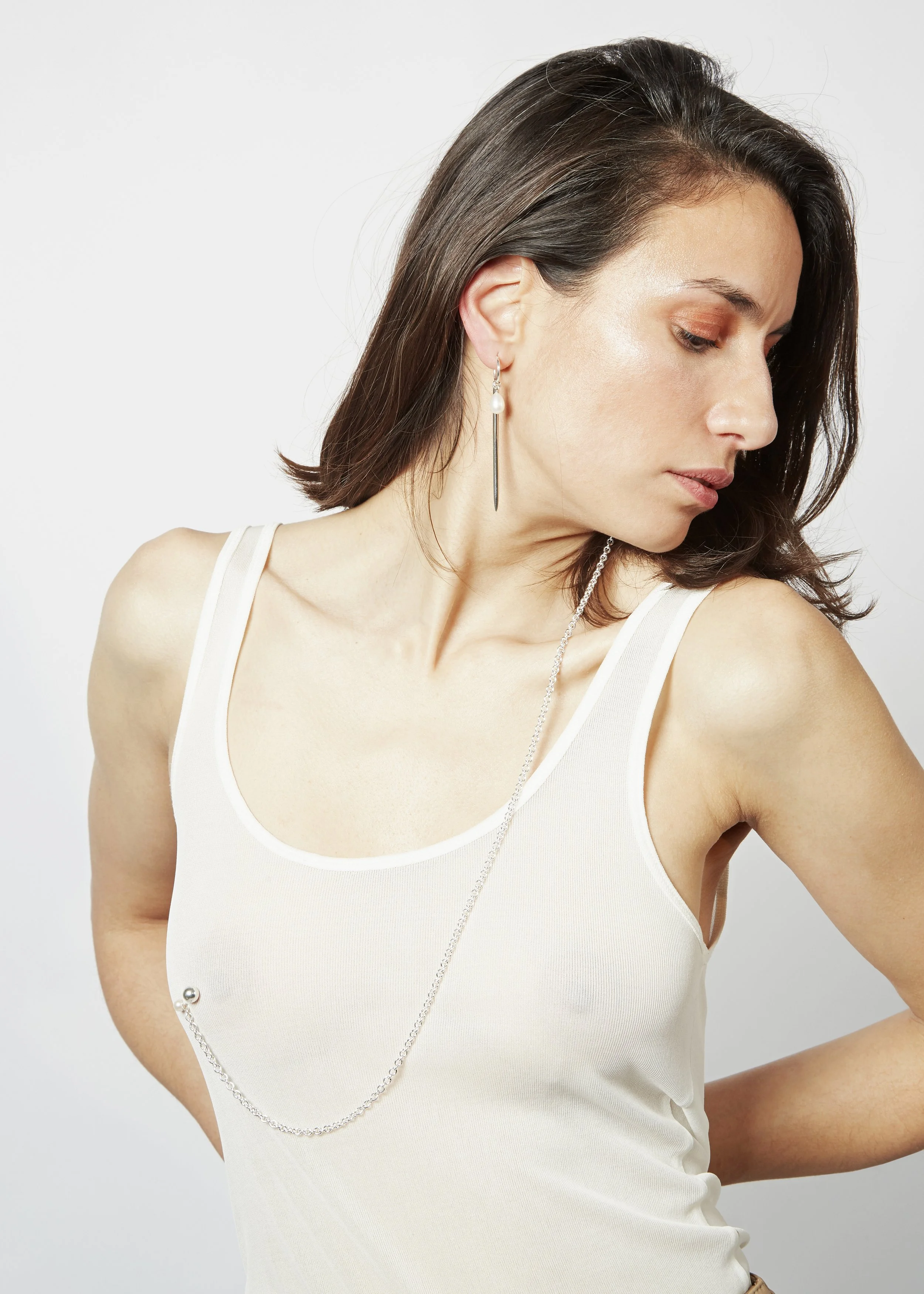 A woman with short dark hair wearing a white sleeveless top, earrings, and a long chain necklace against a plain background.