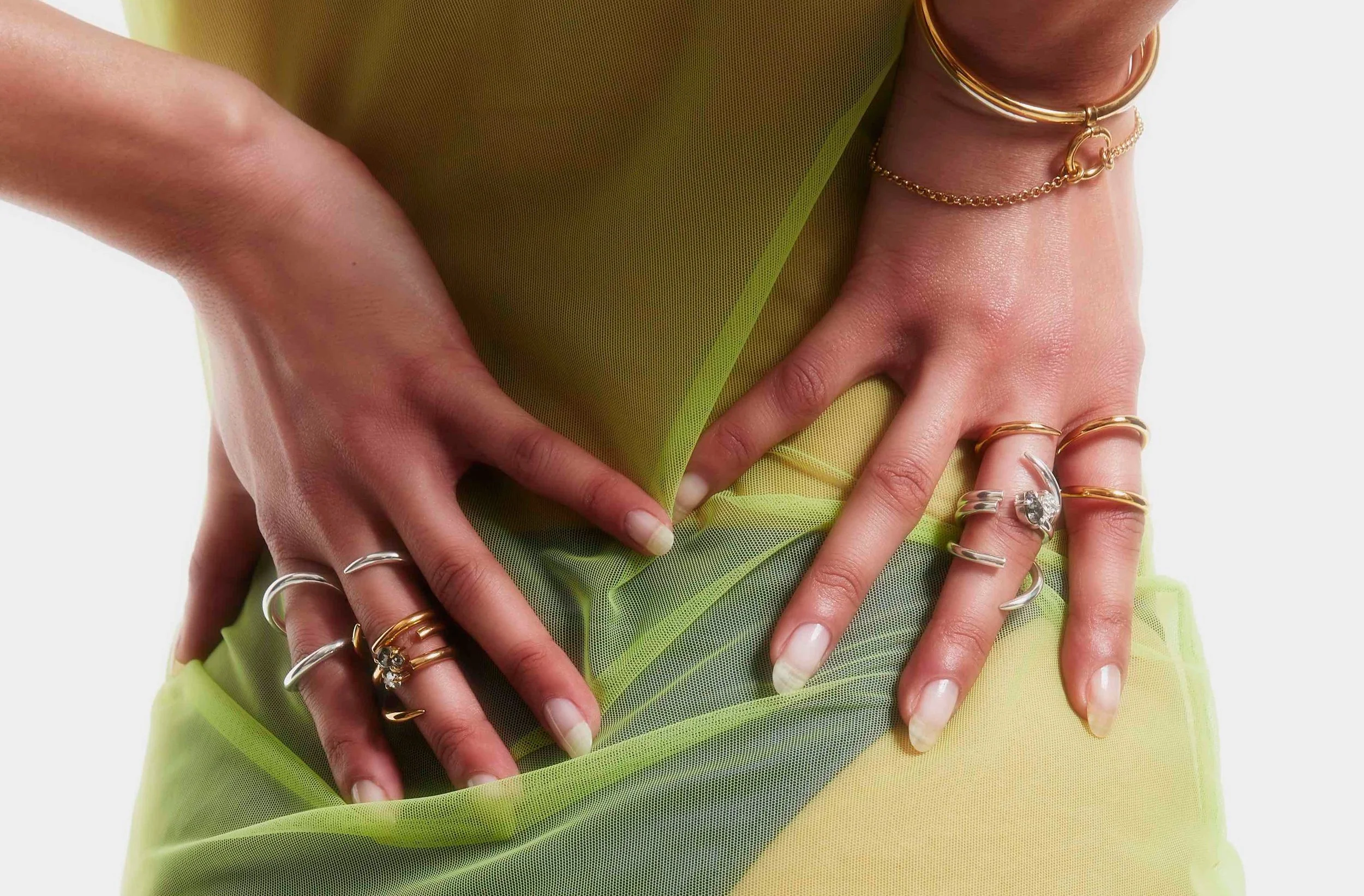 Close-up of a person's hands adorned with multiple rings and bracelets, resting on their waist, wearing a vibrant yellow-green sheer fabric dress.