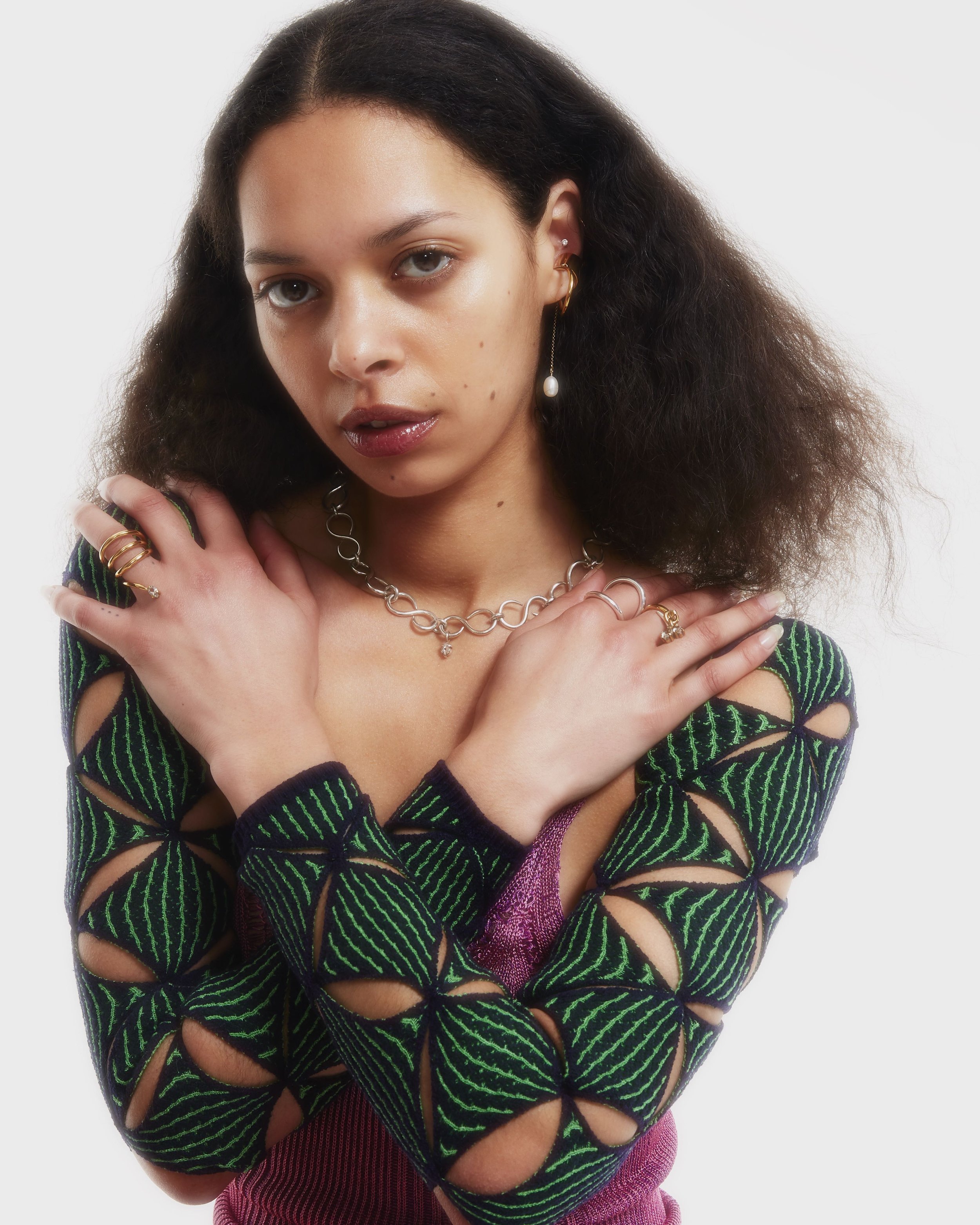 A young woman with curly dark hair wearing a colorful, patterned top with cutouts, multiple rings, a pearl earring, and silver jewelry, posing with her arms crossed over her shoulders against a white background.
