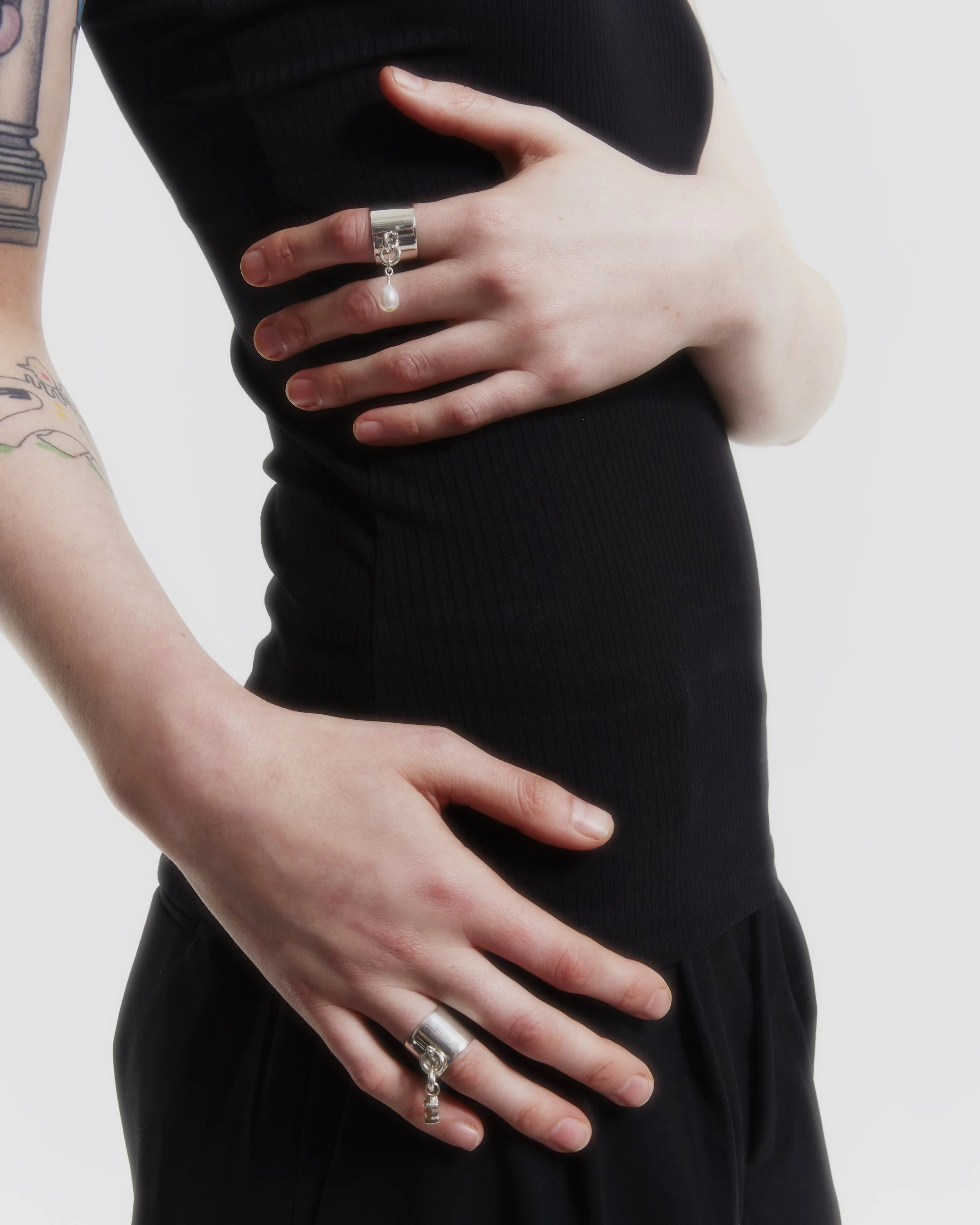 A person wearing a black sleeveless top and black pants. Hands placed on their waist, adorned with rings, one featuring a small pearl charm. Tattooed arms are partially visible.