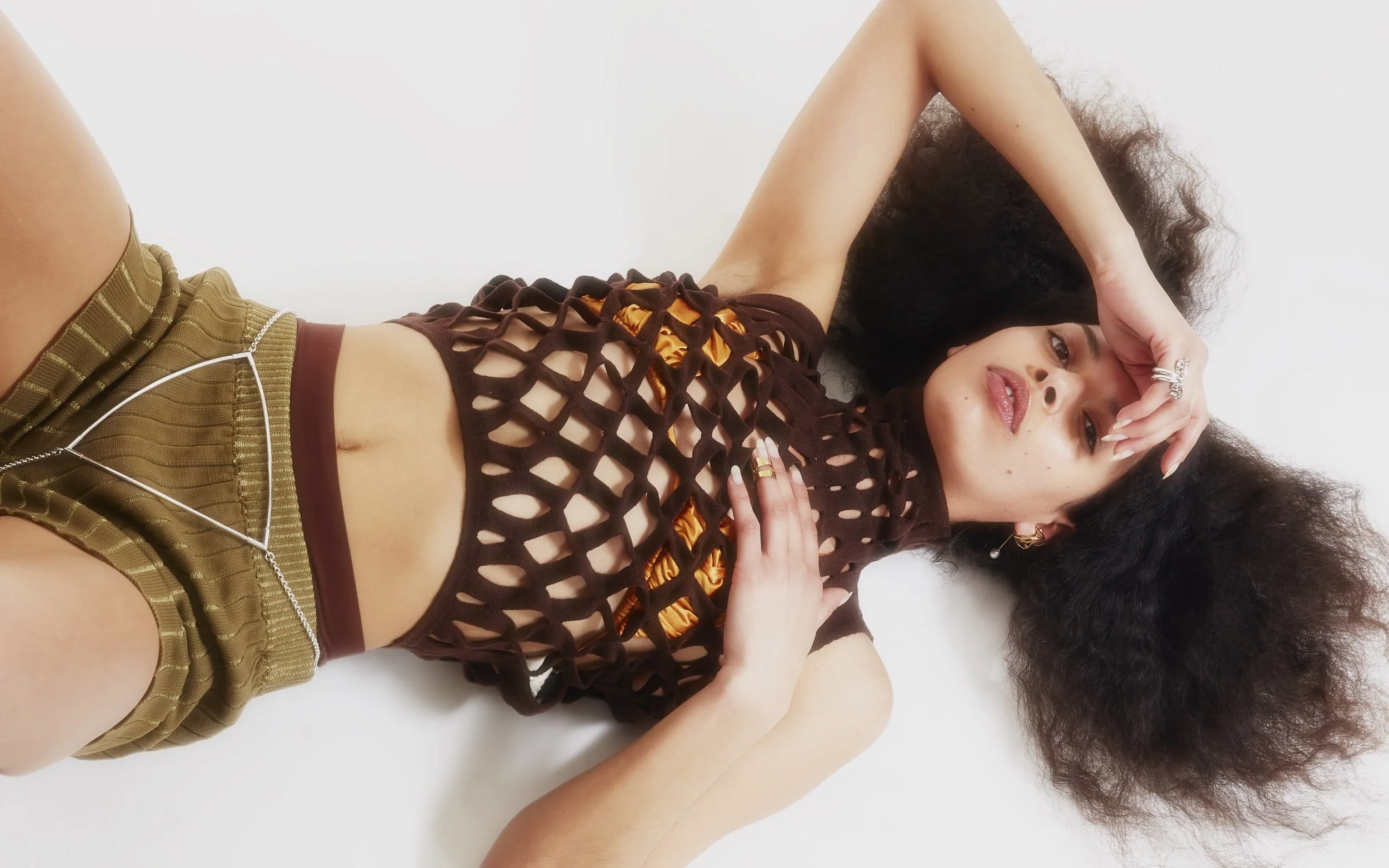 Young woman with curly hair wearing a black fishnet top over a yellow bra and brown shorts, lying on a white surface with one hand on her forehead and the other on her chest, looking at the camera.