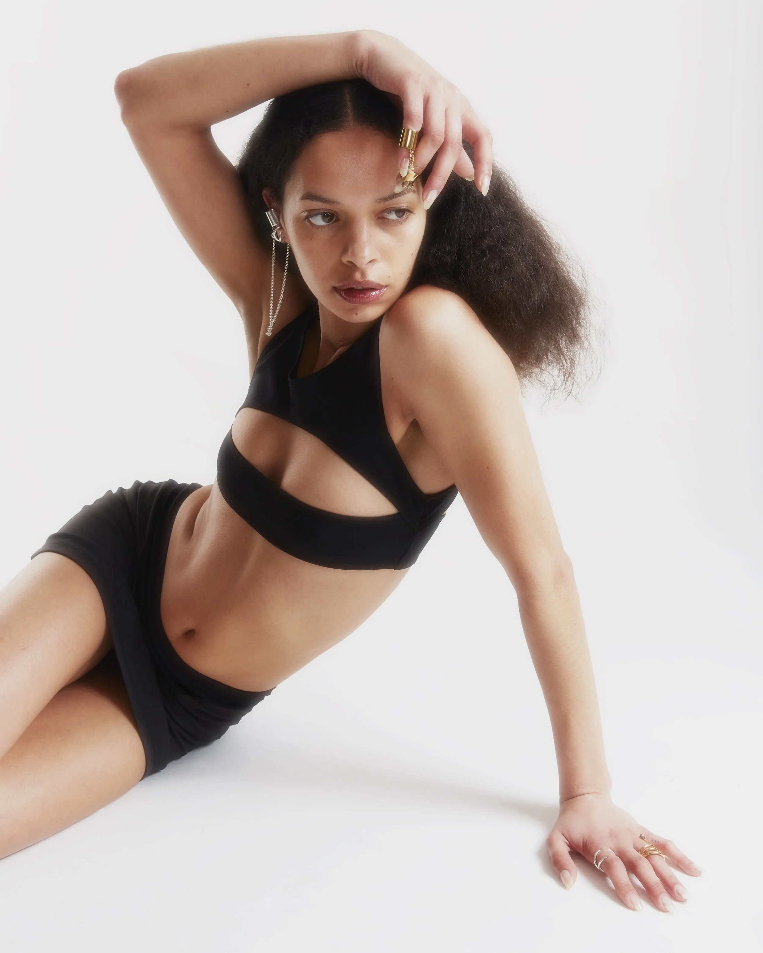 A woman with curly hair poses against a plain white background, wearing a black cutout top and shorts, with one arm resting on the floor and the other raised above her head.