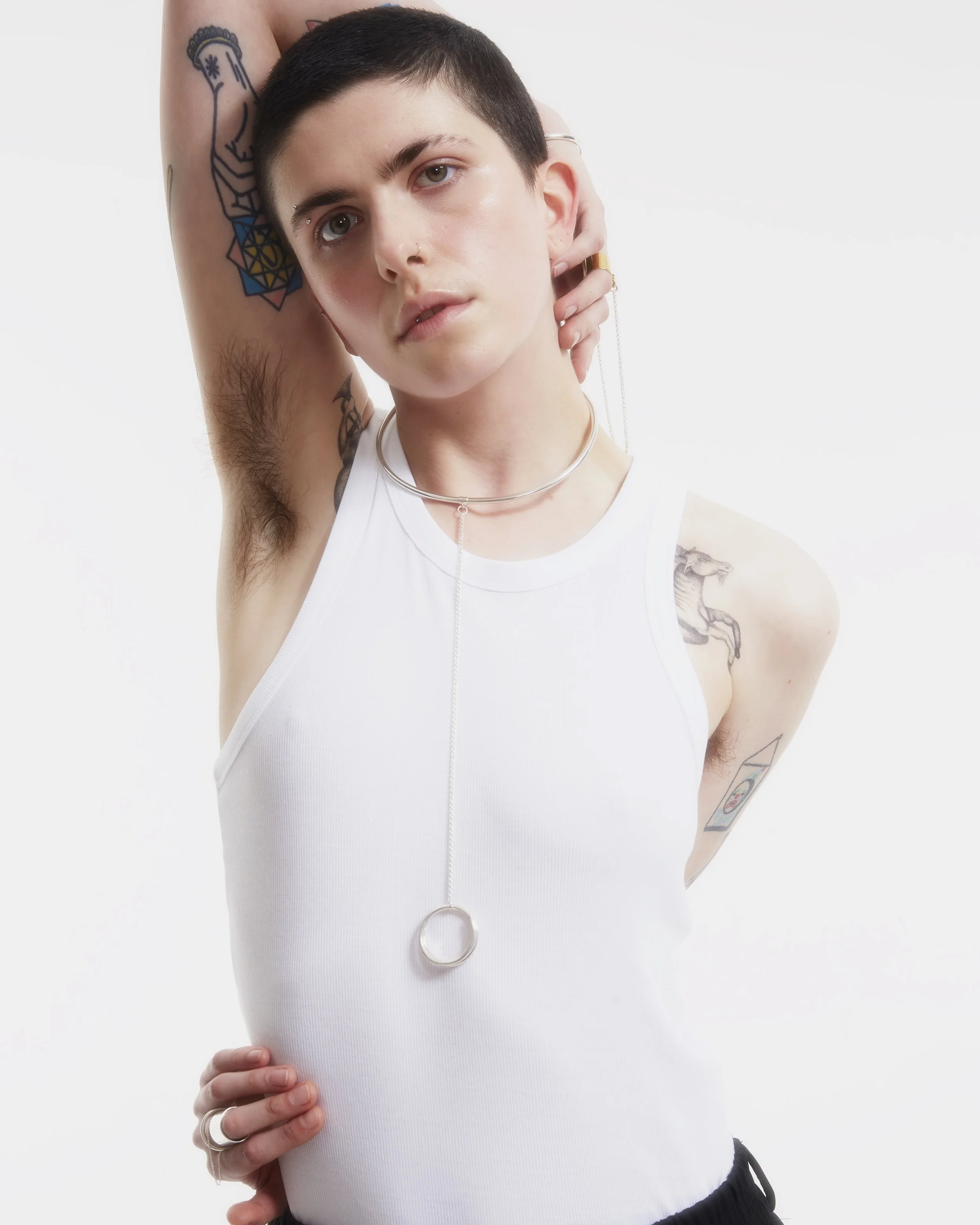 Person with short dark hair, tattoos, and piercings posing with one arm lifted, wearing a white sleeveless top and jewelry, against a plain white background.